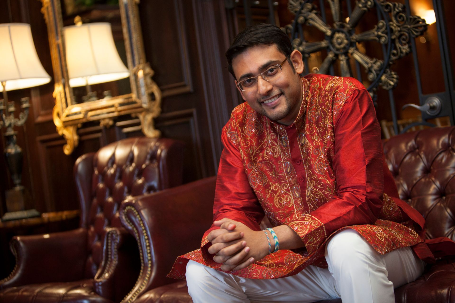 Man in red embroidered kurta sitting on a leather chair in a wood-paneled room with warm lamps.
