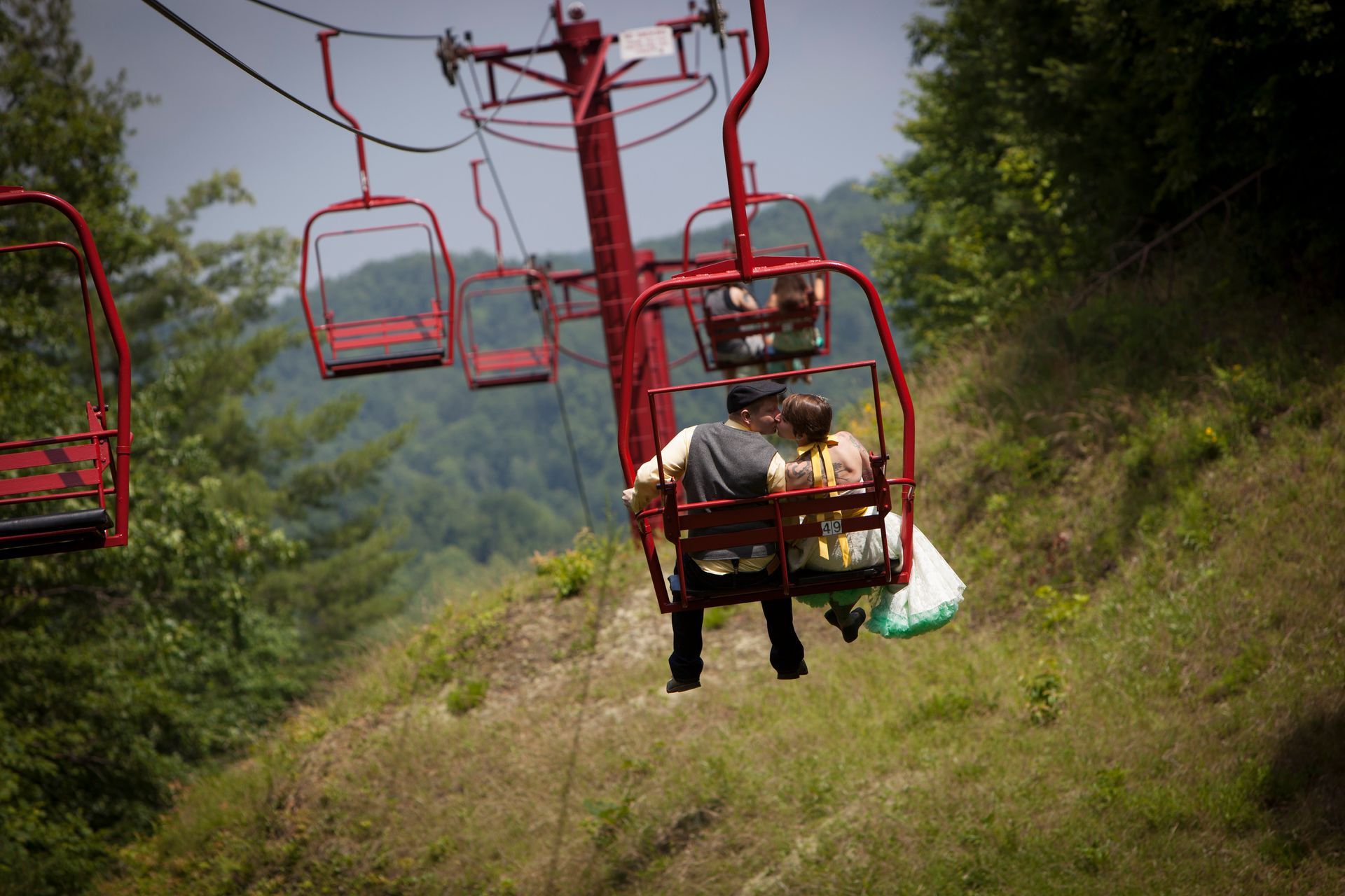 Red ski lift chairs carrying two people over a green hillside with trees and mountains in the distance