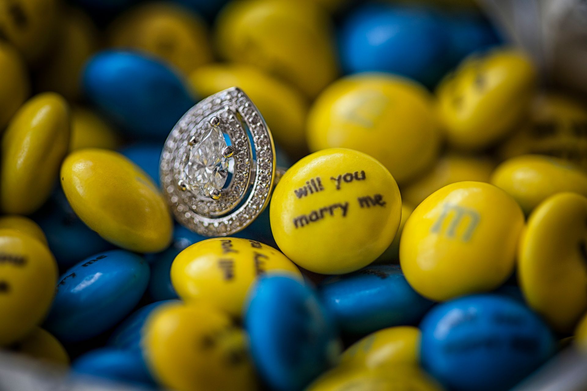 Diamond engagement ring with “Will you marry me?” on a yellow candy among blue and yellow candies
