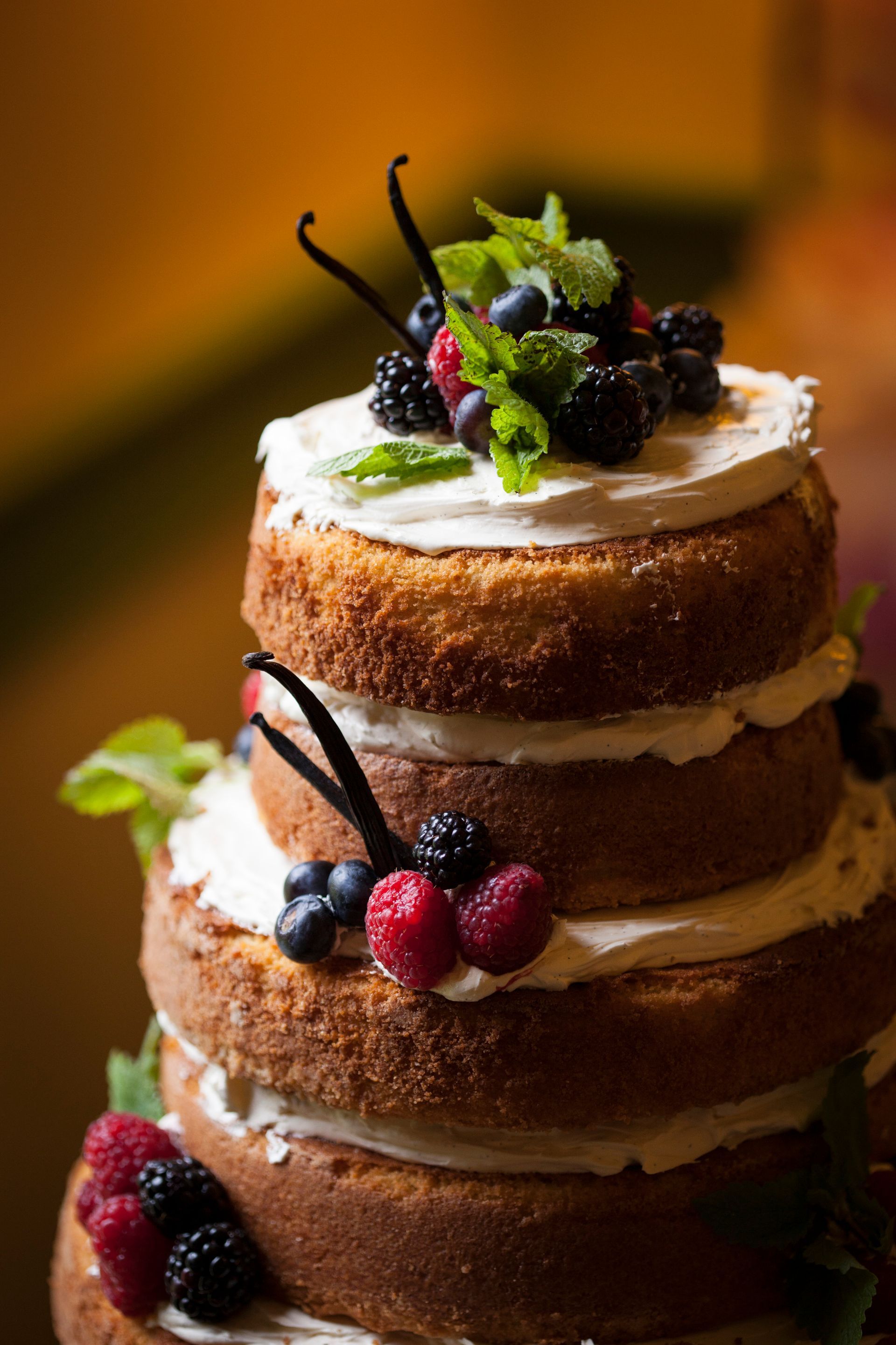 Stacked naked cake with white frosting, blackberries, raspberries, mint, and vanilla pods on top.