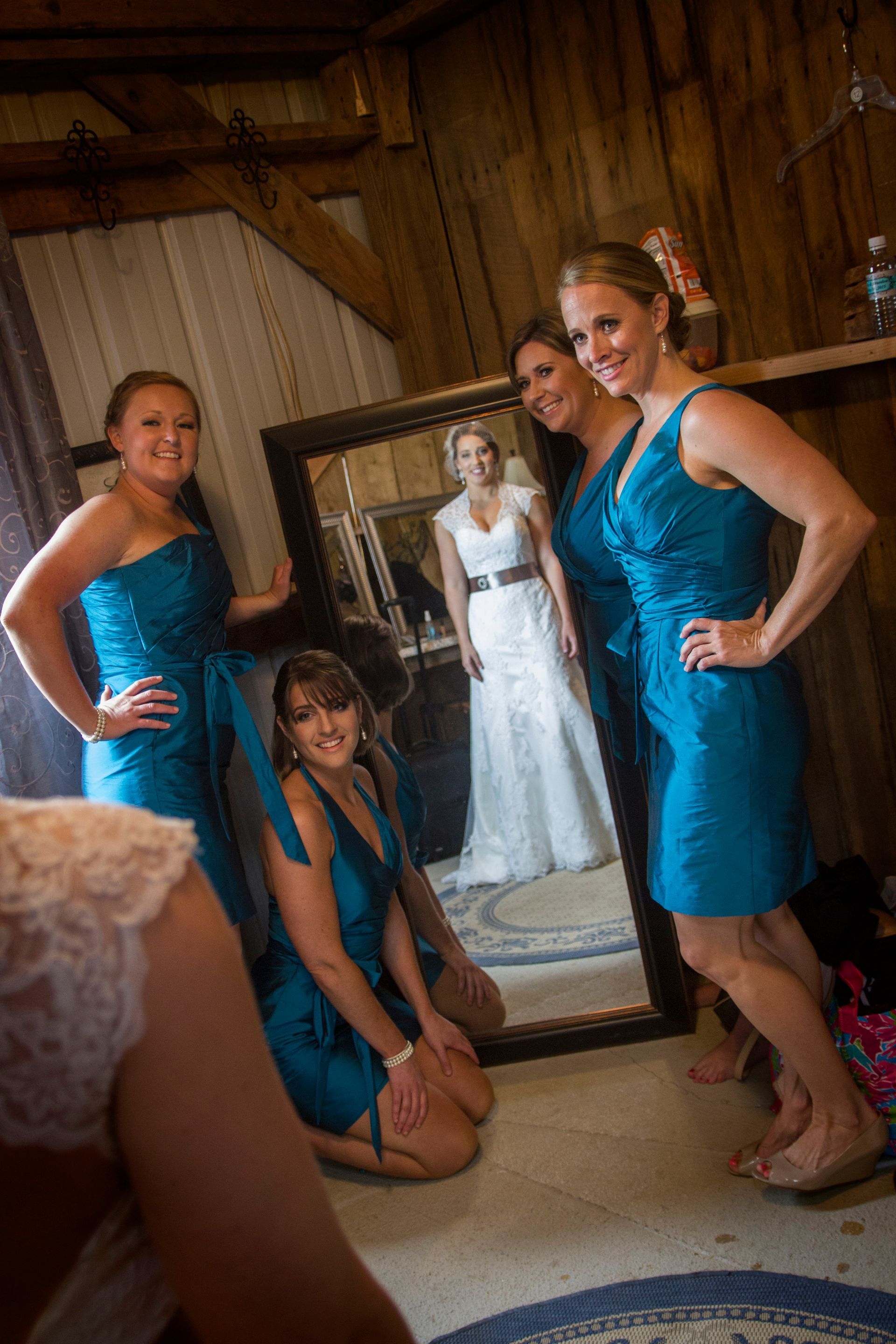 Bridesmaids in blue dresses pose around a mirror reflecting a white wedding gown indoors