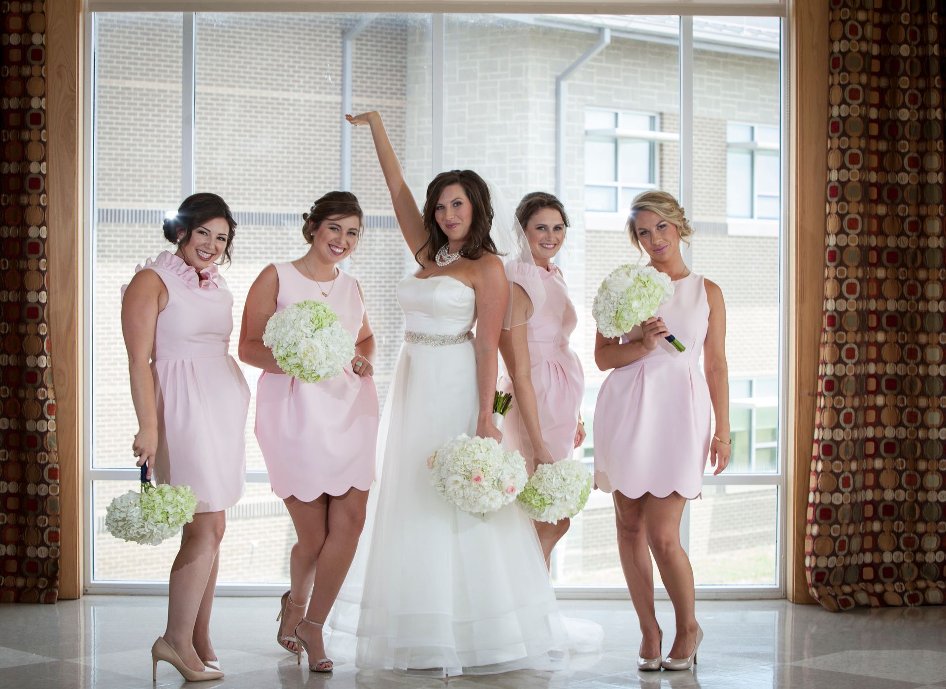 Bride and four bridesmaids in pink dresses posing indoors, one holding a bouquet aloft.