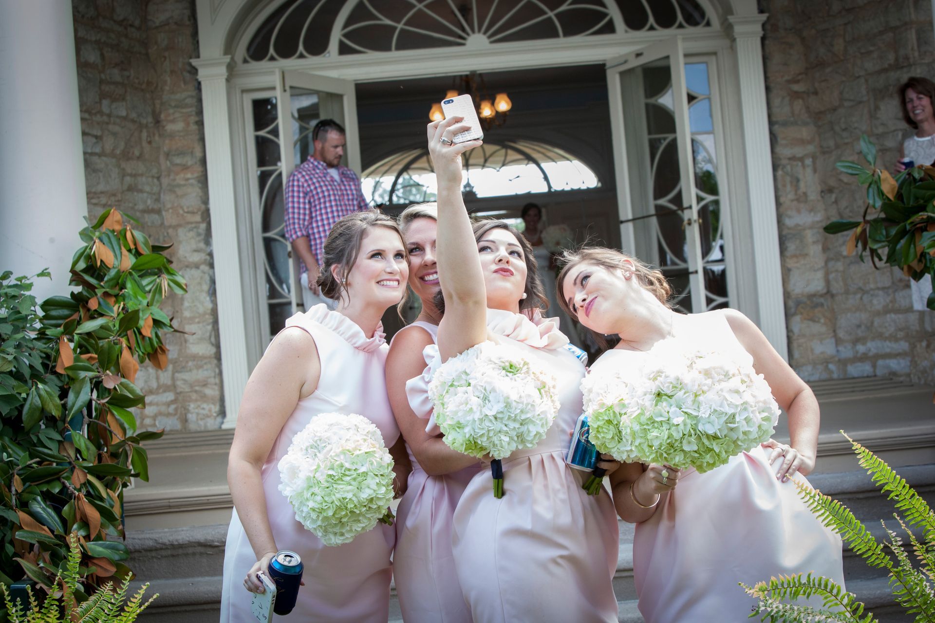 Three women in lavender dresses pose with white bouquets outside a stone house, one raising a bouquet overhead.