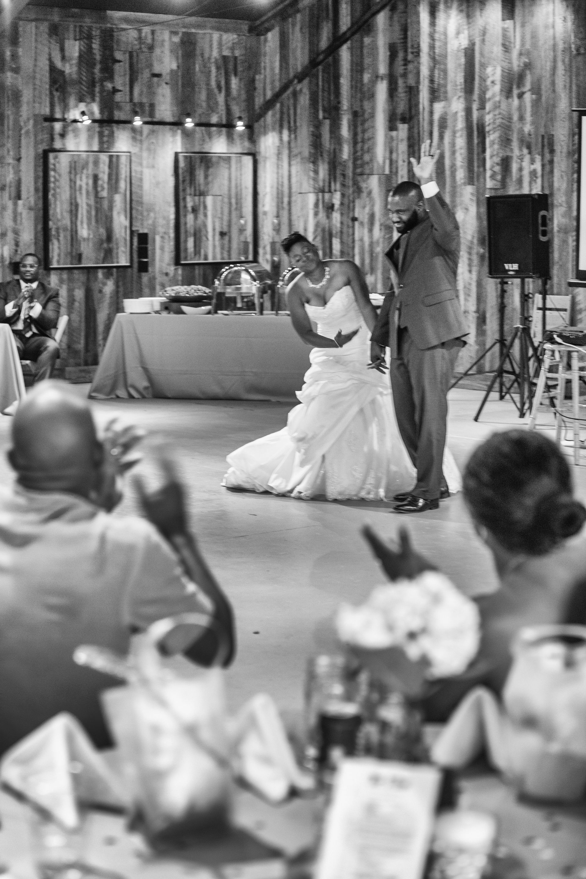 Bride kneels beside groom at a rustic wedding reception, guests watching in the foreground