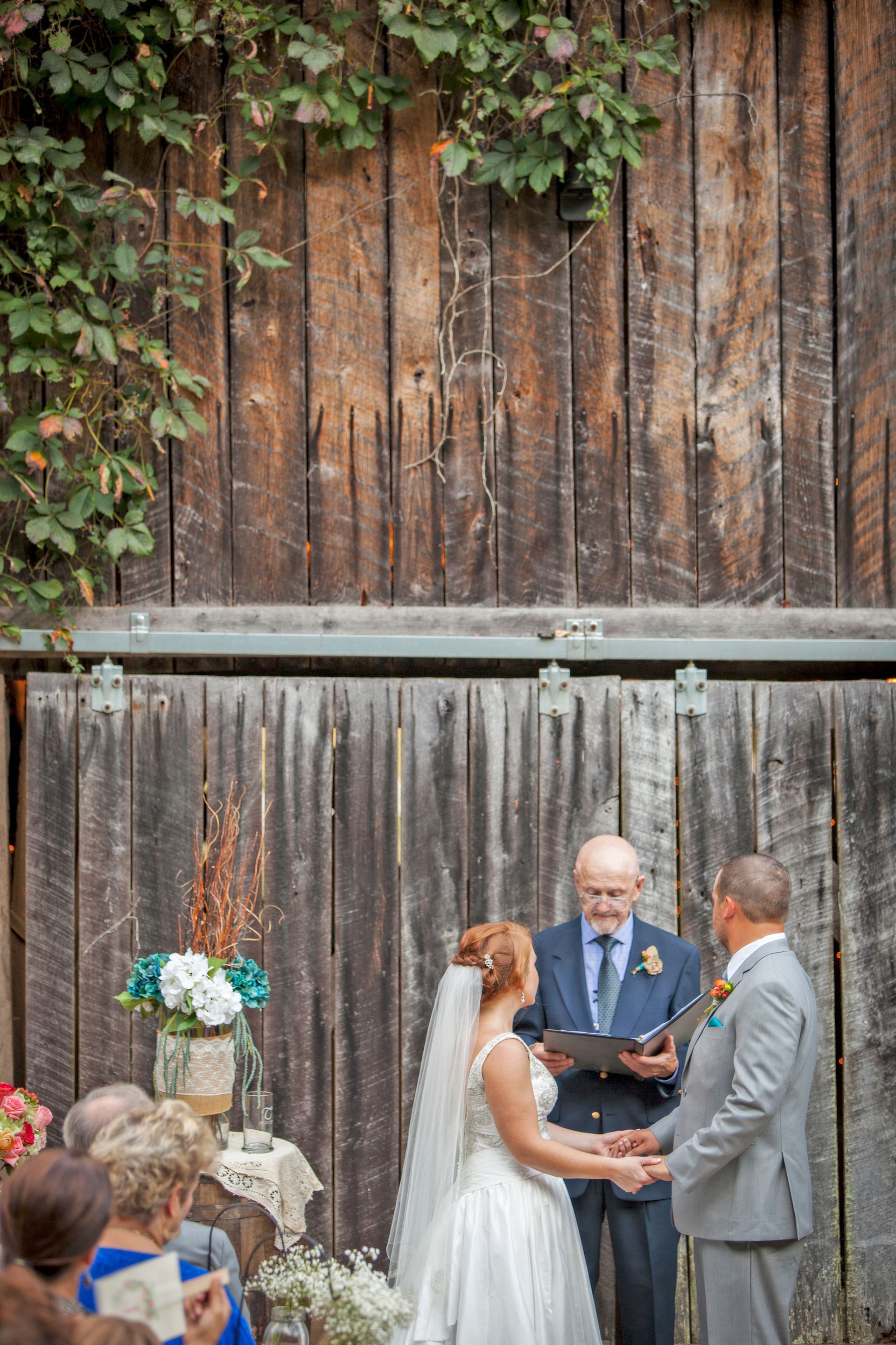 Wedding ceremony outdoors before a rustic wooden backdrop, couple exchanging vows with officiant nearby