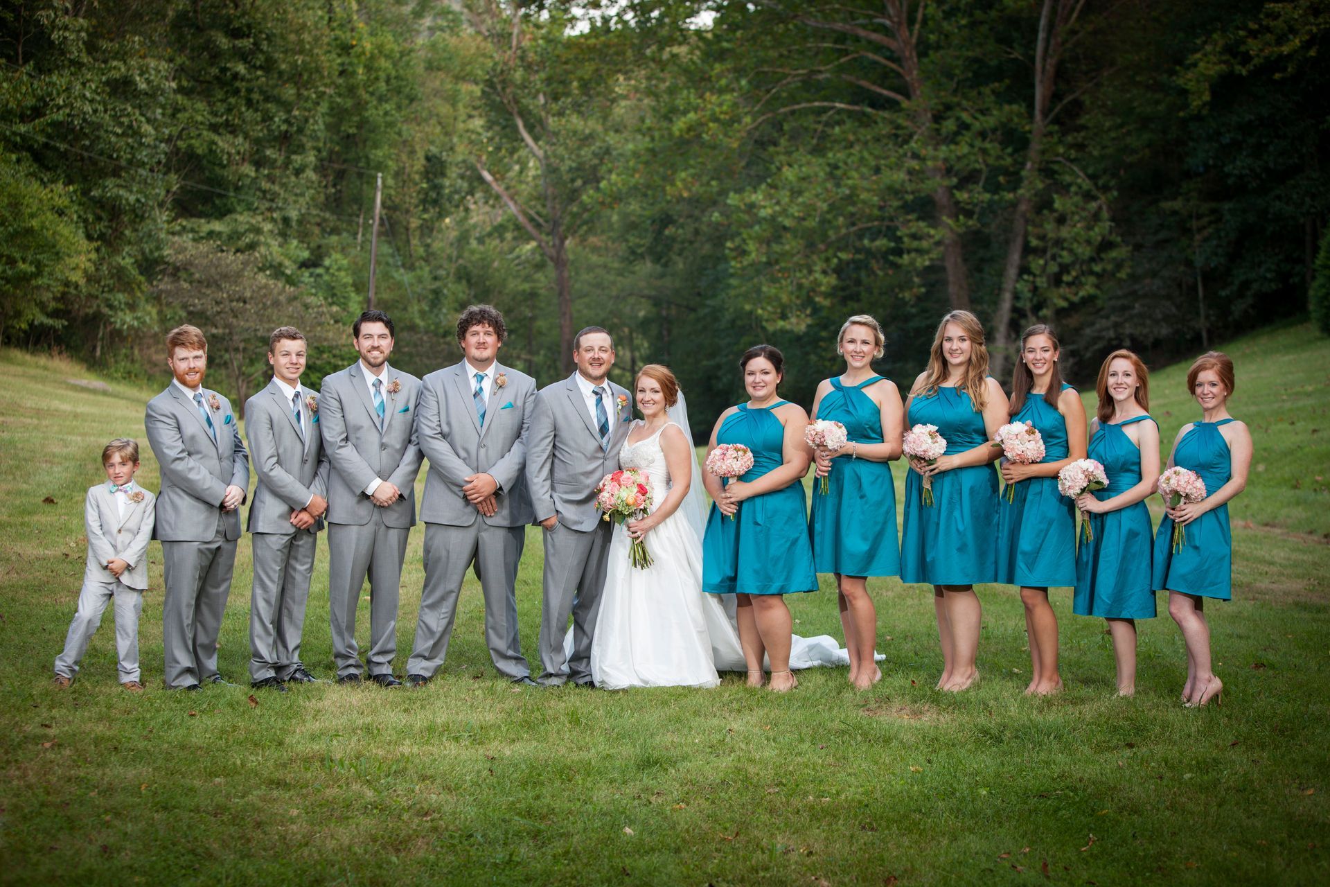 Wedding party posing outdoors in a grassy field, with the bride in white and attendants in gray and teal.