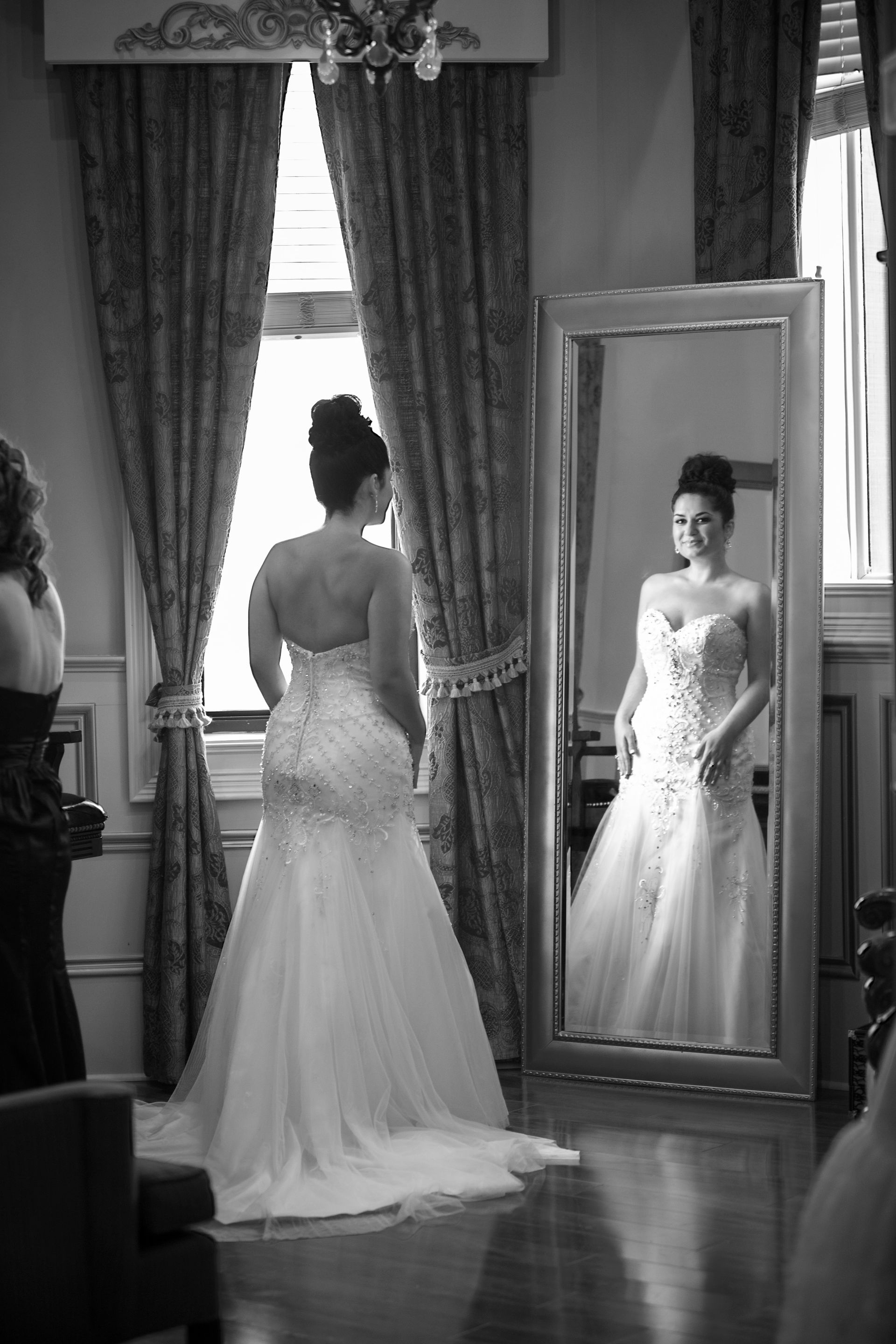 Bride in a strapless wedding gown looking at her reflection in a full-length mirror inside a formal room.