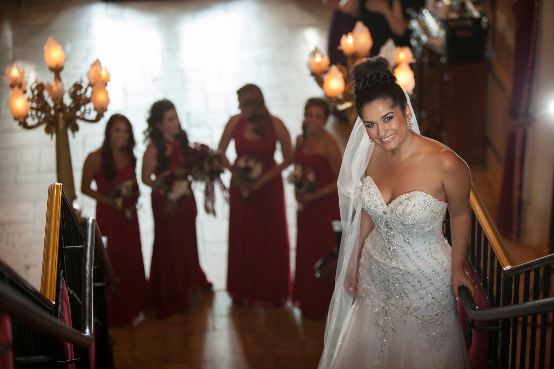 Bride in a white gown descends a staircase, with bridesmaids in red dresses behind her.