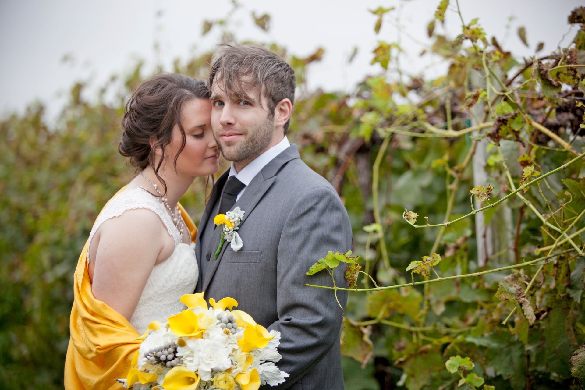 Couple in wedding attire posing outdoors in a vineyard, with yellow bouquet and flowers