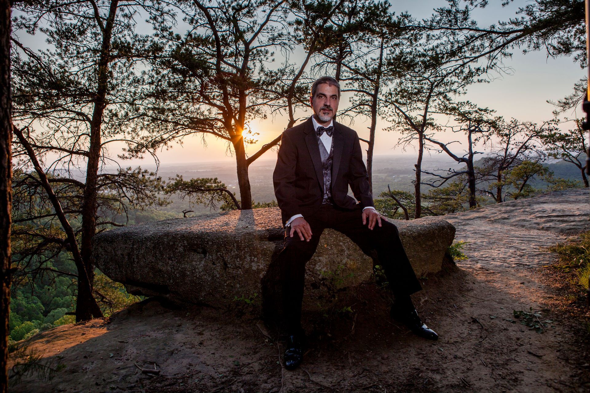 Man in a suit sits on a stone bench at sunset in a wooded park.