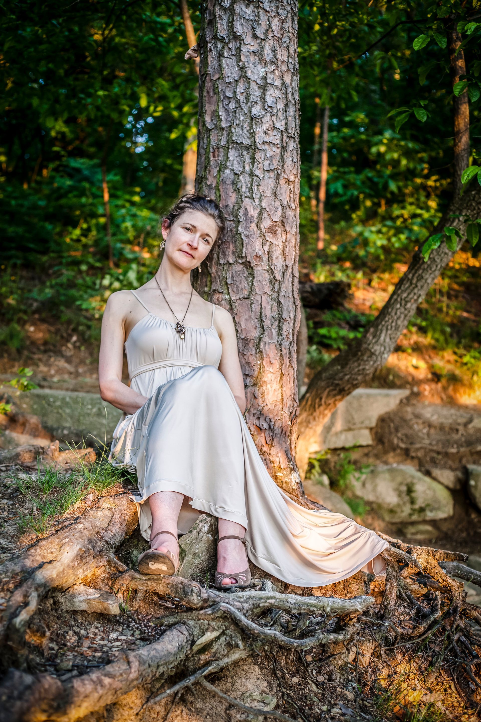 Woman in a light dress sitting by a tree on exposed roots in a wooded outdoor setting