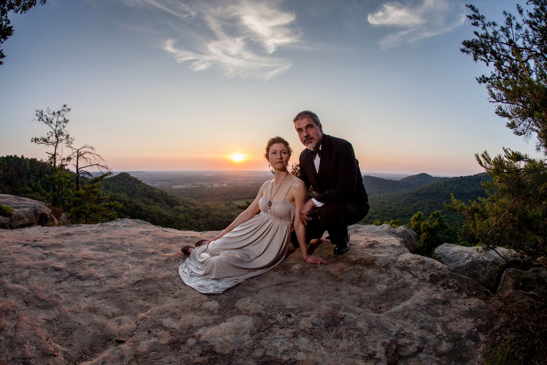 Couple seated on rocky overlook at sunset, with a sweeping valley and dramatic sky behind them