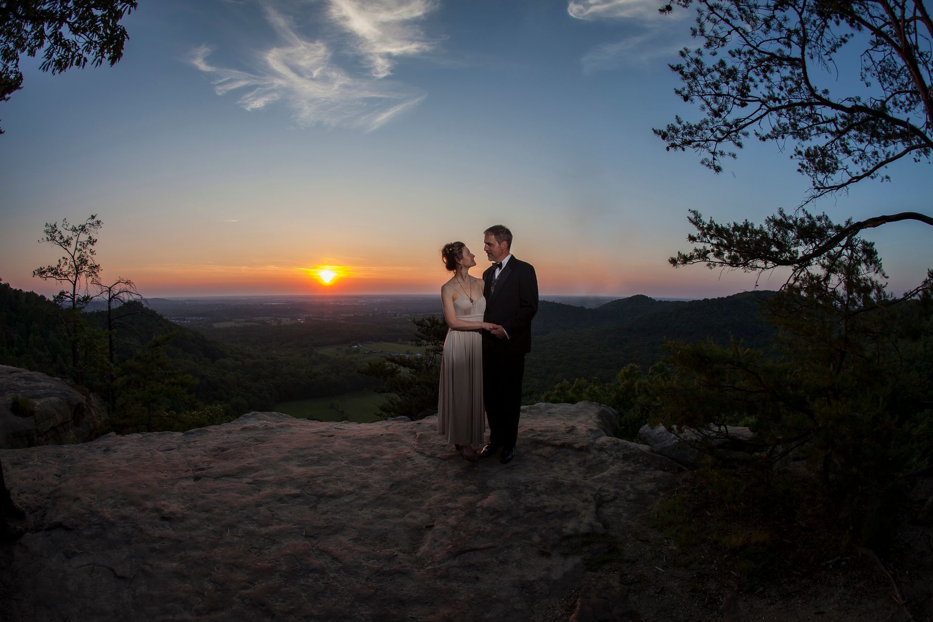 Couple in formal attire standing at sunset on a rocky overlook with a glowing horizon