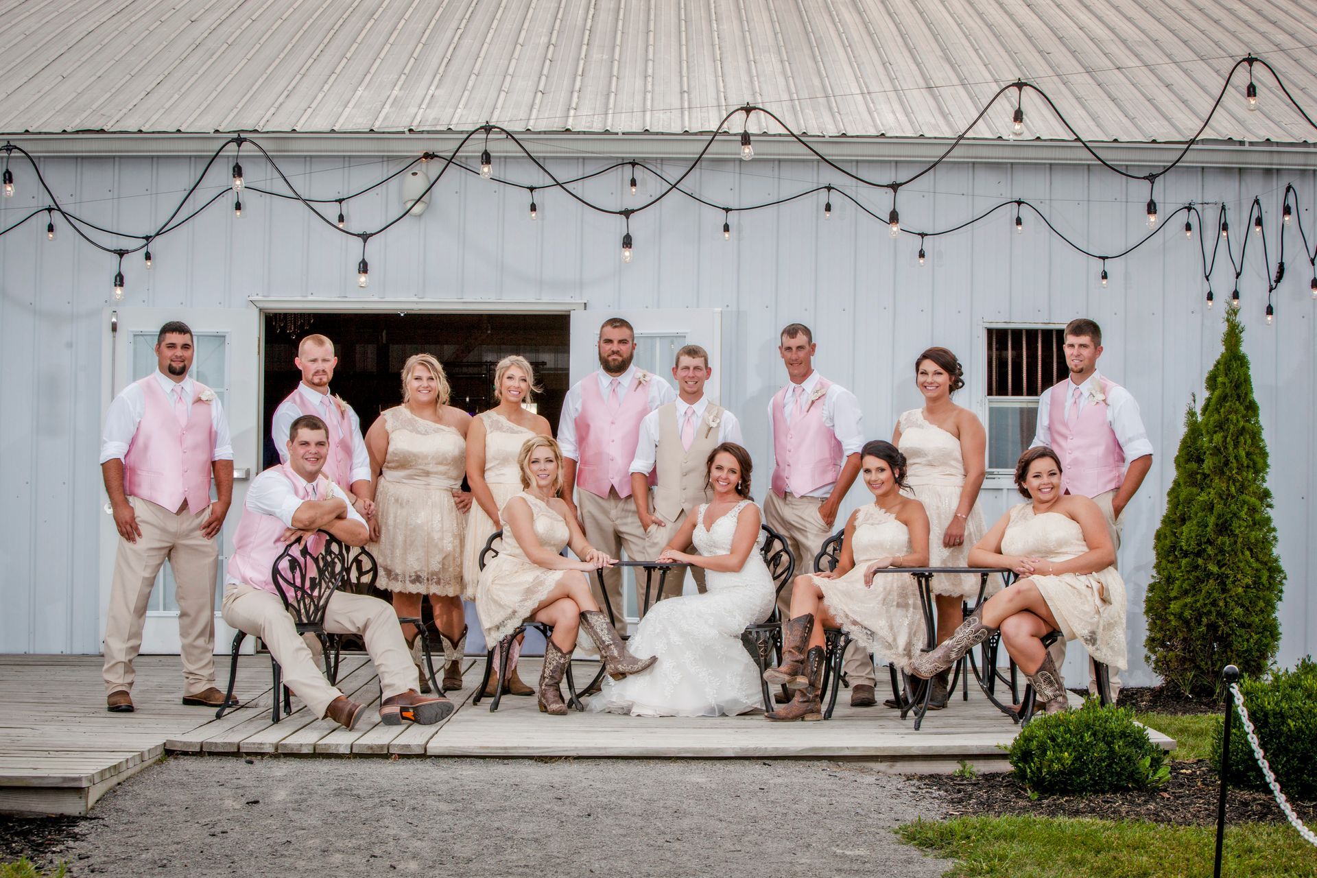 Wedding party posing outdoors in front of a white barn with string lights