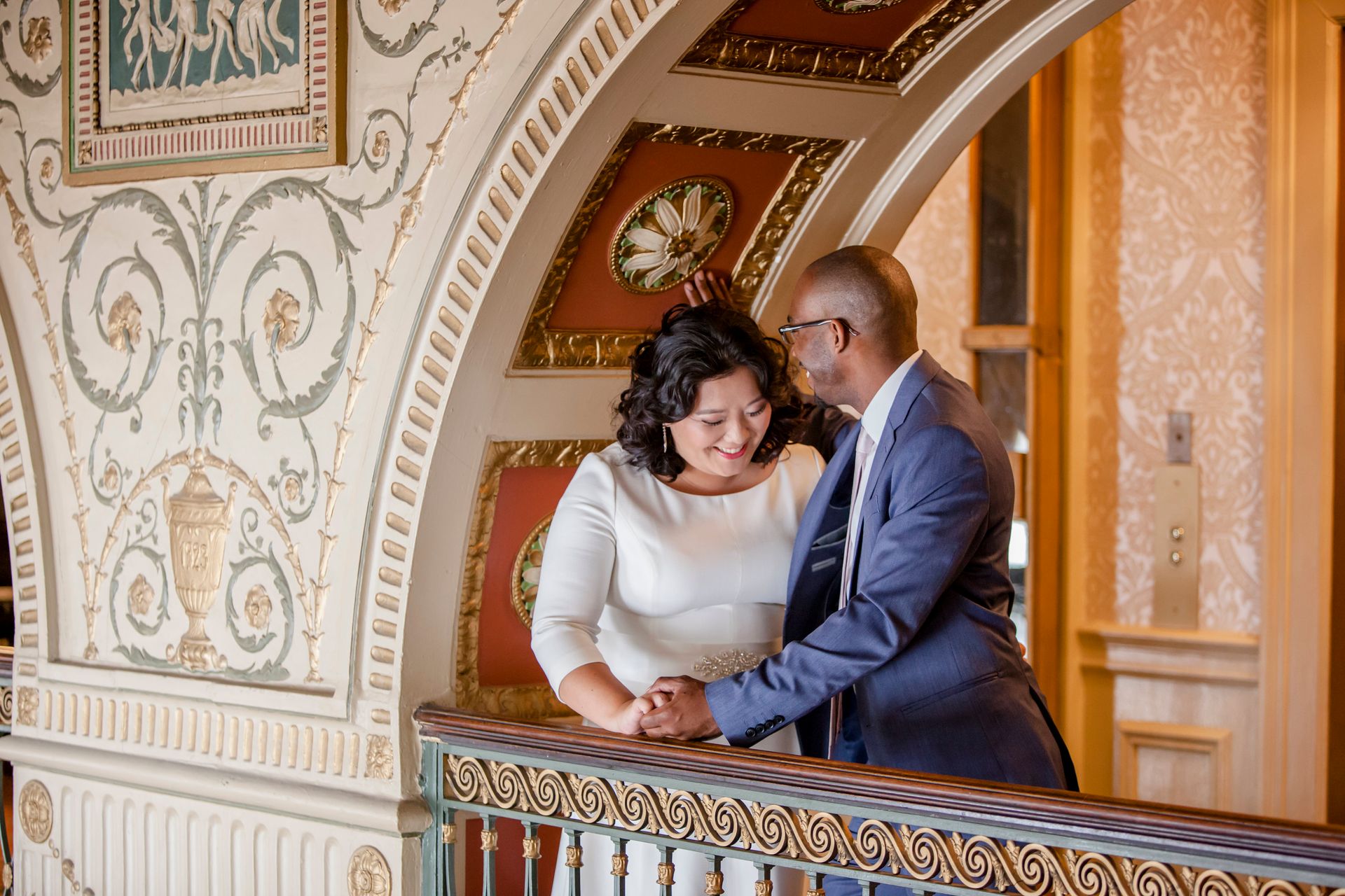 Two people smiling on an ornate balcony, leaning together in a richly decorated interior.