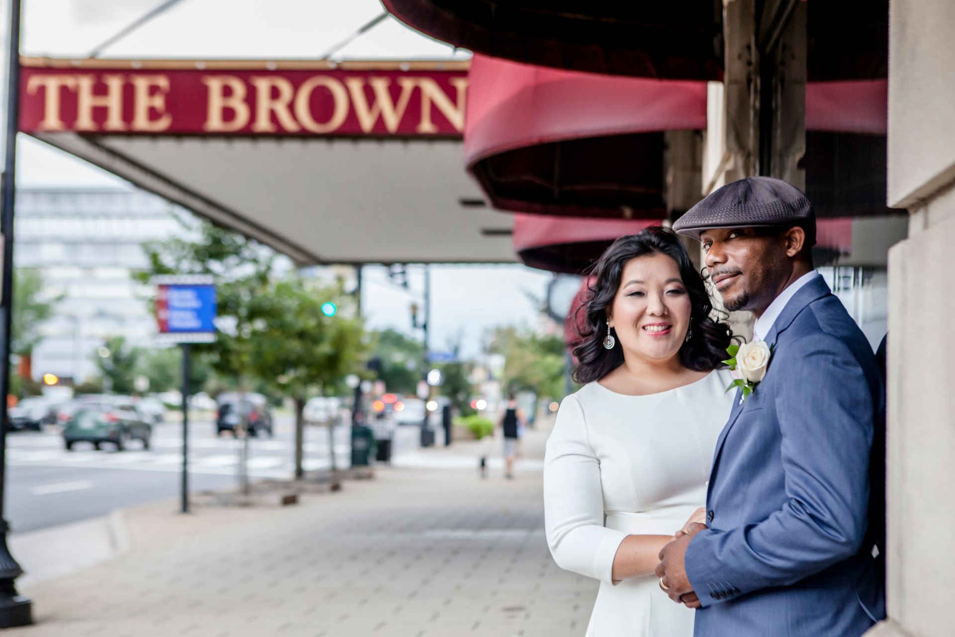 Smiling couple in formal attire standing under a red awning on a city sidewalk.