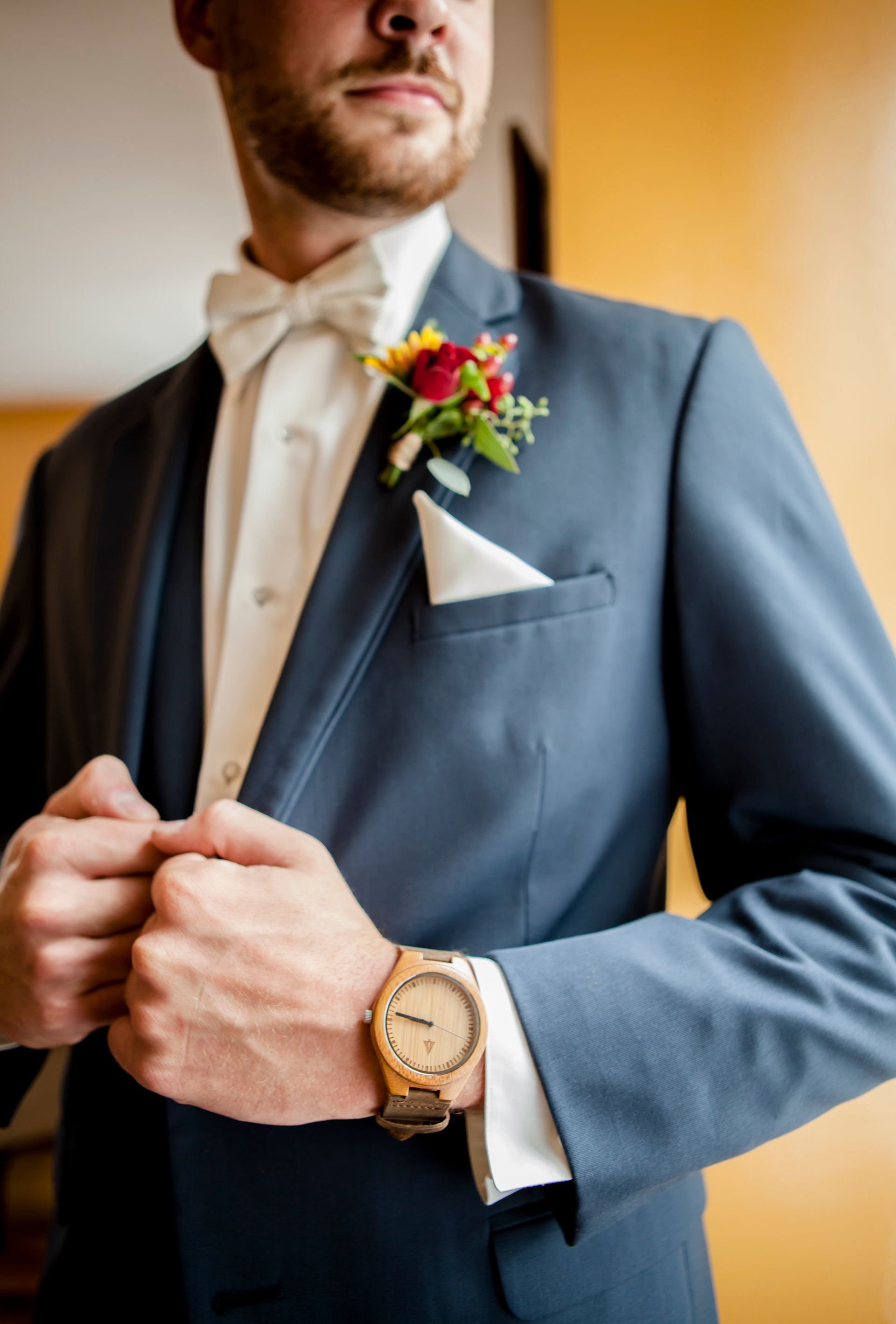 Man in a blue suit adjusting his jacket, with a white shirt, boutonniere, and gold watch