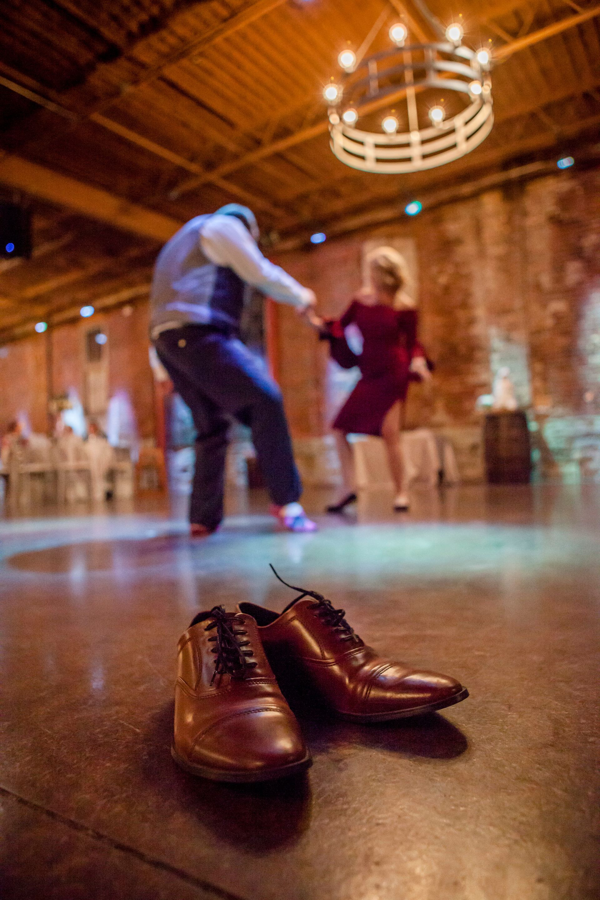 Brown dress shoes in the foreground, with two people dancing in a warmly lit rustic hall behind them