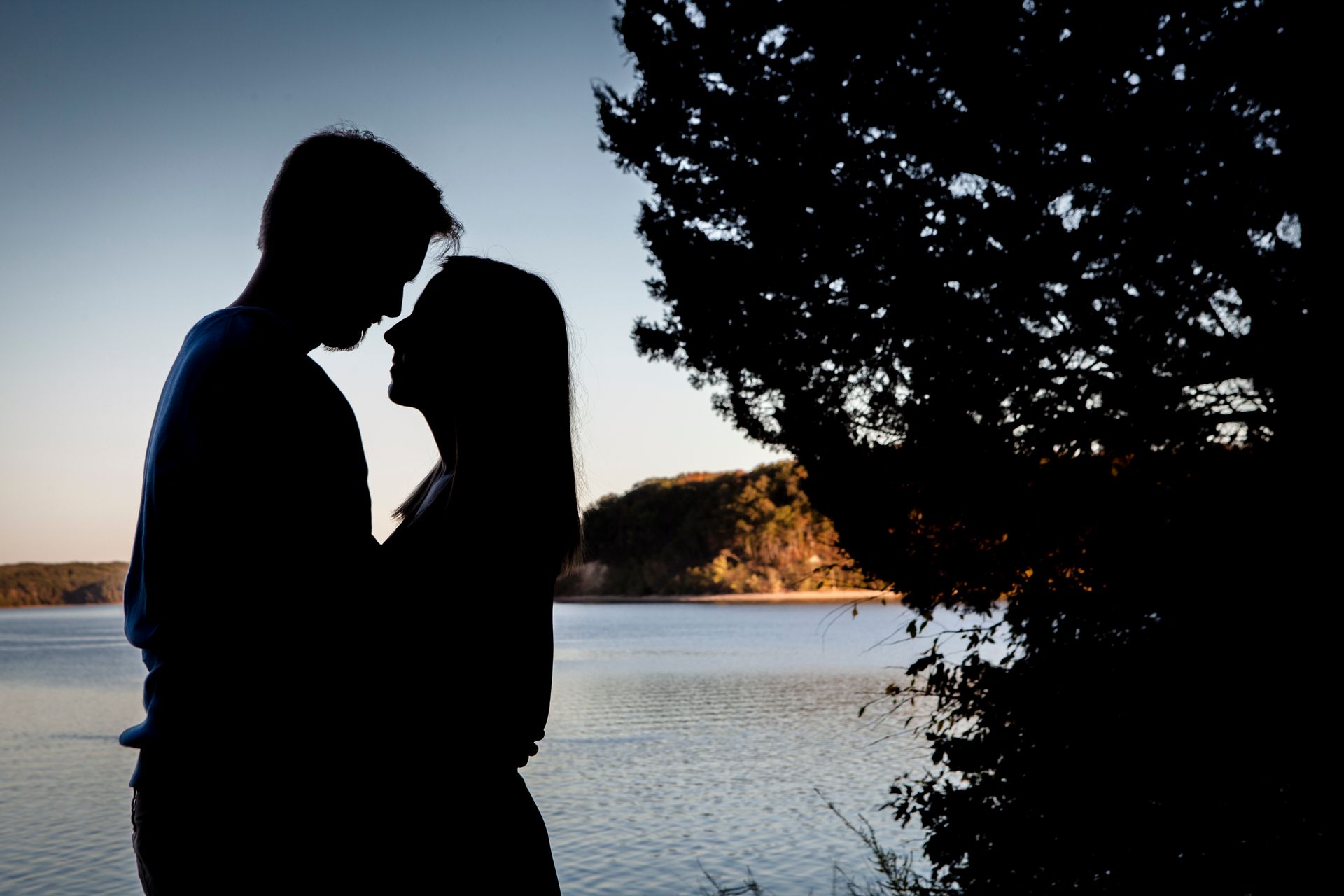 Silhouetted couple embracing by a lakeshore at sunset, framed by trees.