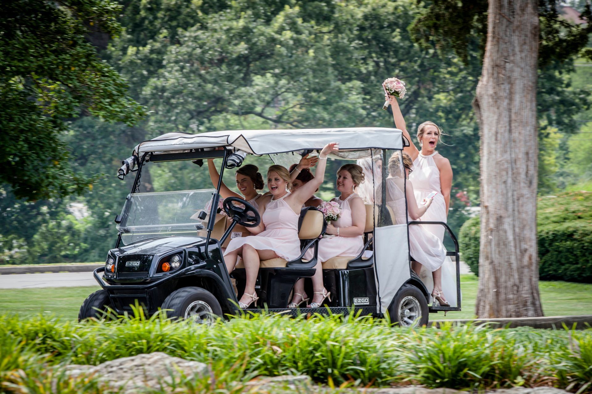 People riding in a golf cart on a tree-lined path, one waving with a trophy raised.