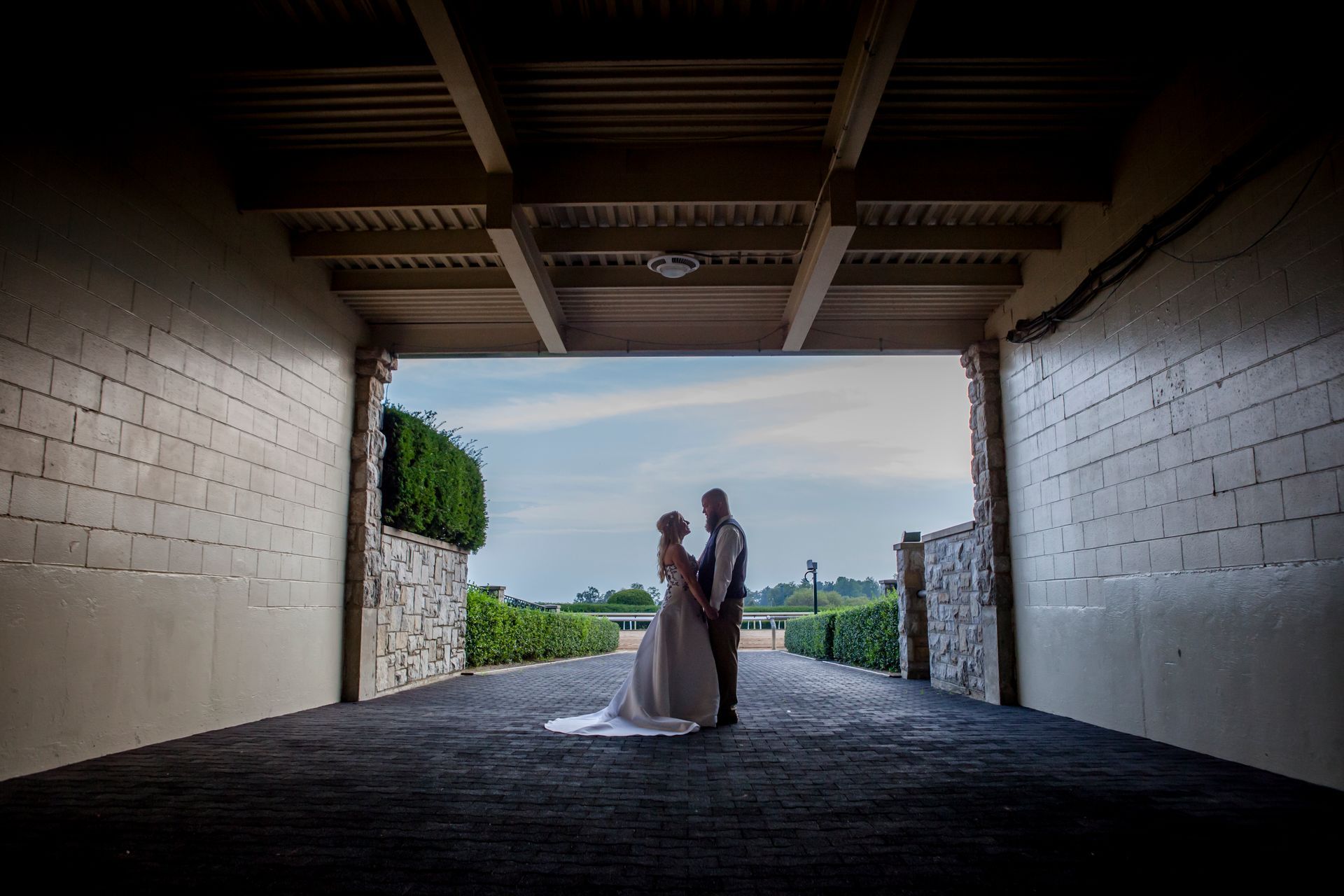 Bride and groom standing under a tunnel, silhouetted against daylight at an outdoor venue.