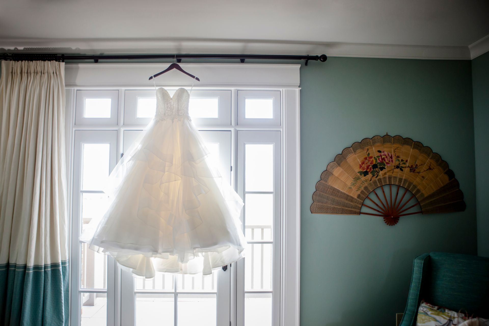 White dress hanging in a sunlit window beside a decorative fan on a teal wall.