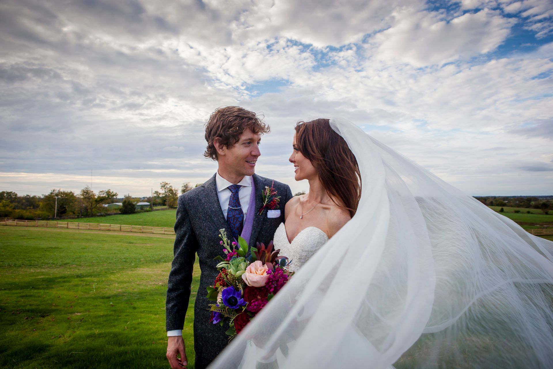 Bride and groom standing in a grassy field, smiling under a cloudy sky, with the veil flowing in the wind