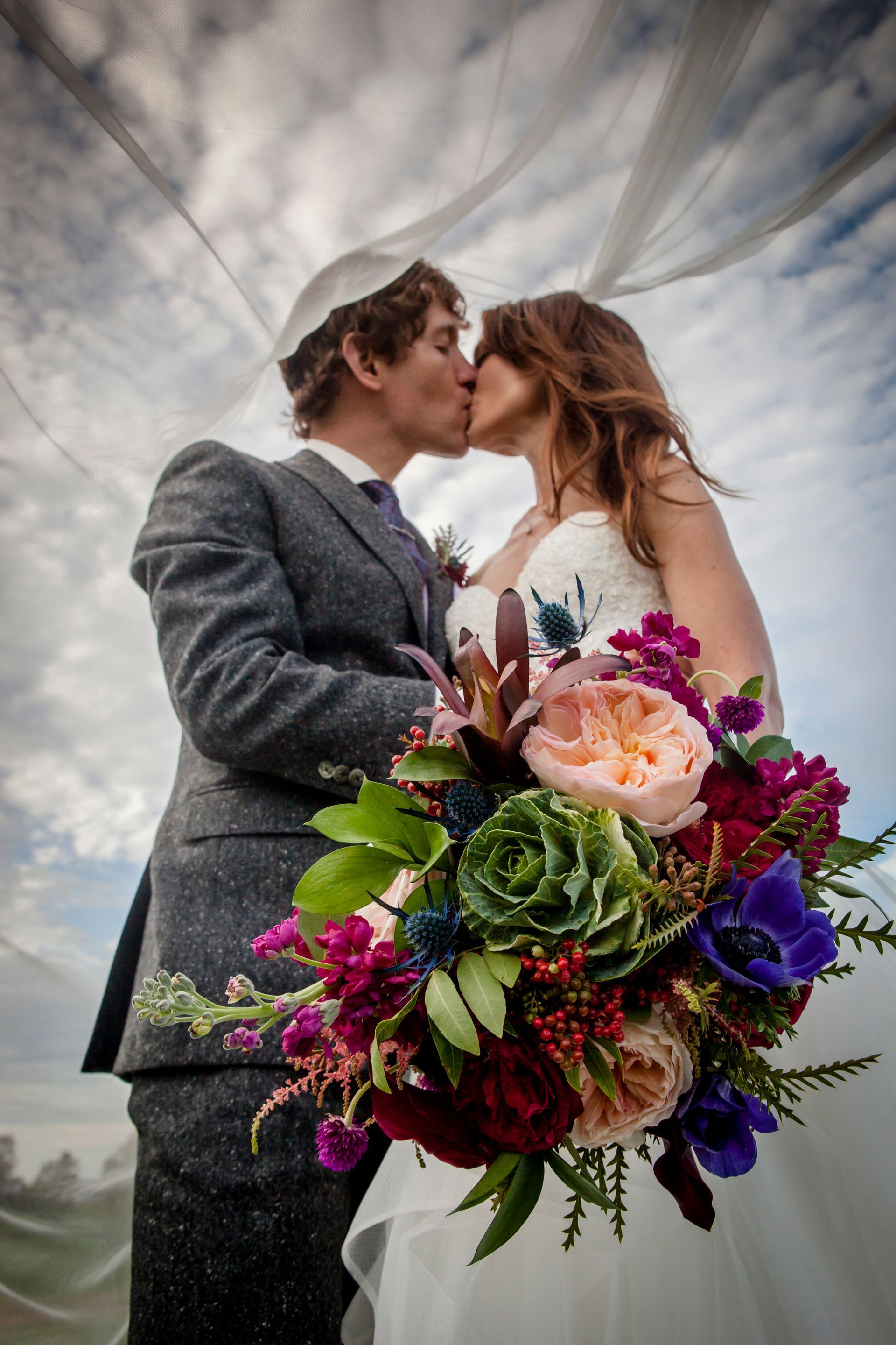 Wedding couple kissing under a billowing veil, with a colorful bouquet in the foreground