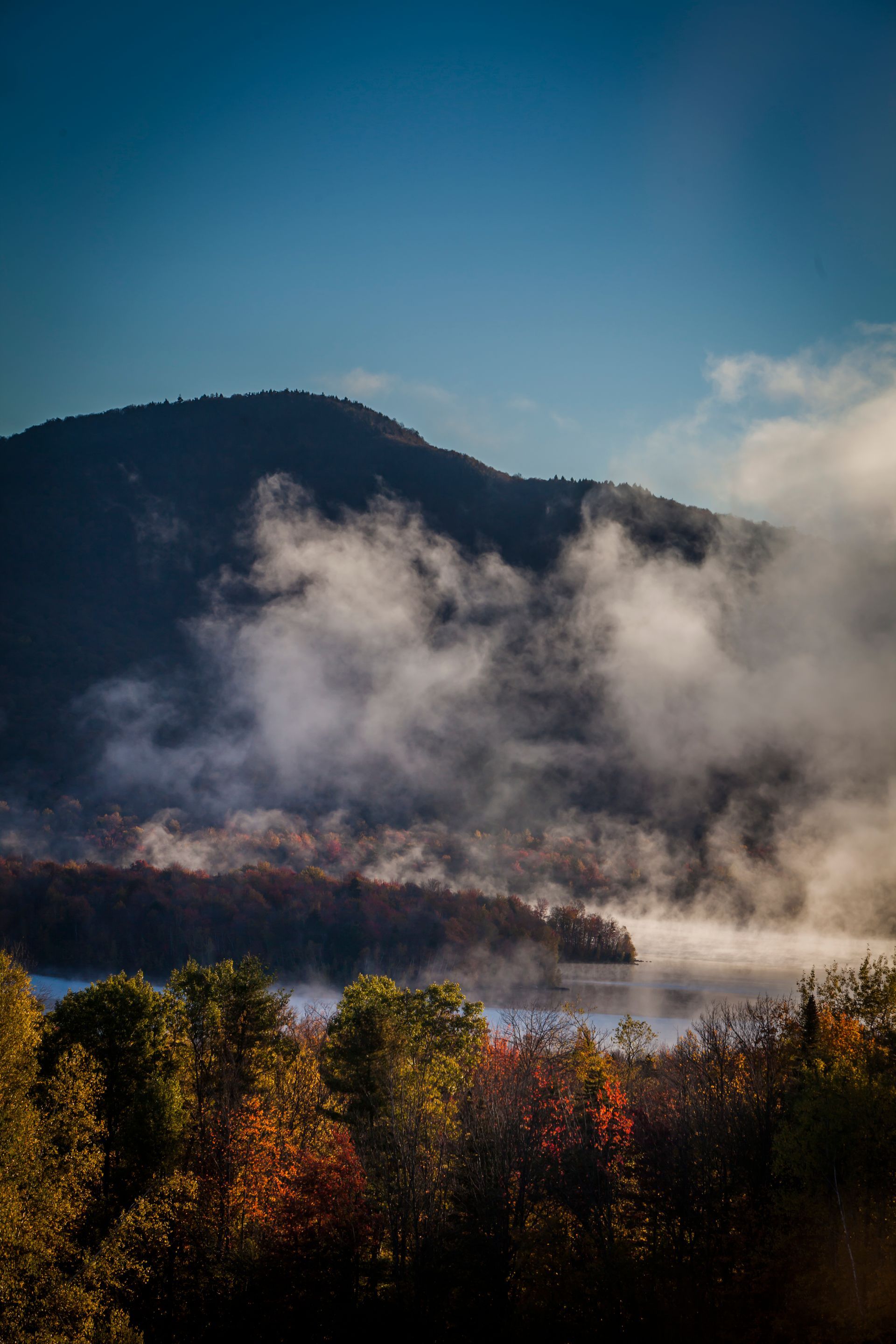 Misty mountain lake at sunrise with clouds, dark hills, and autumn-colored trees in the foreground