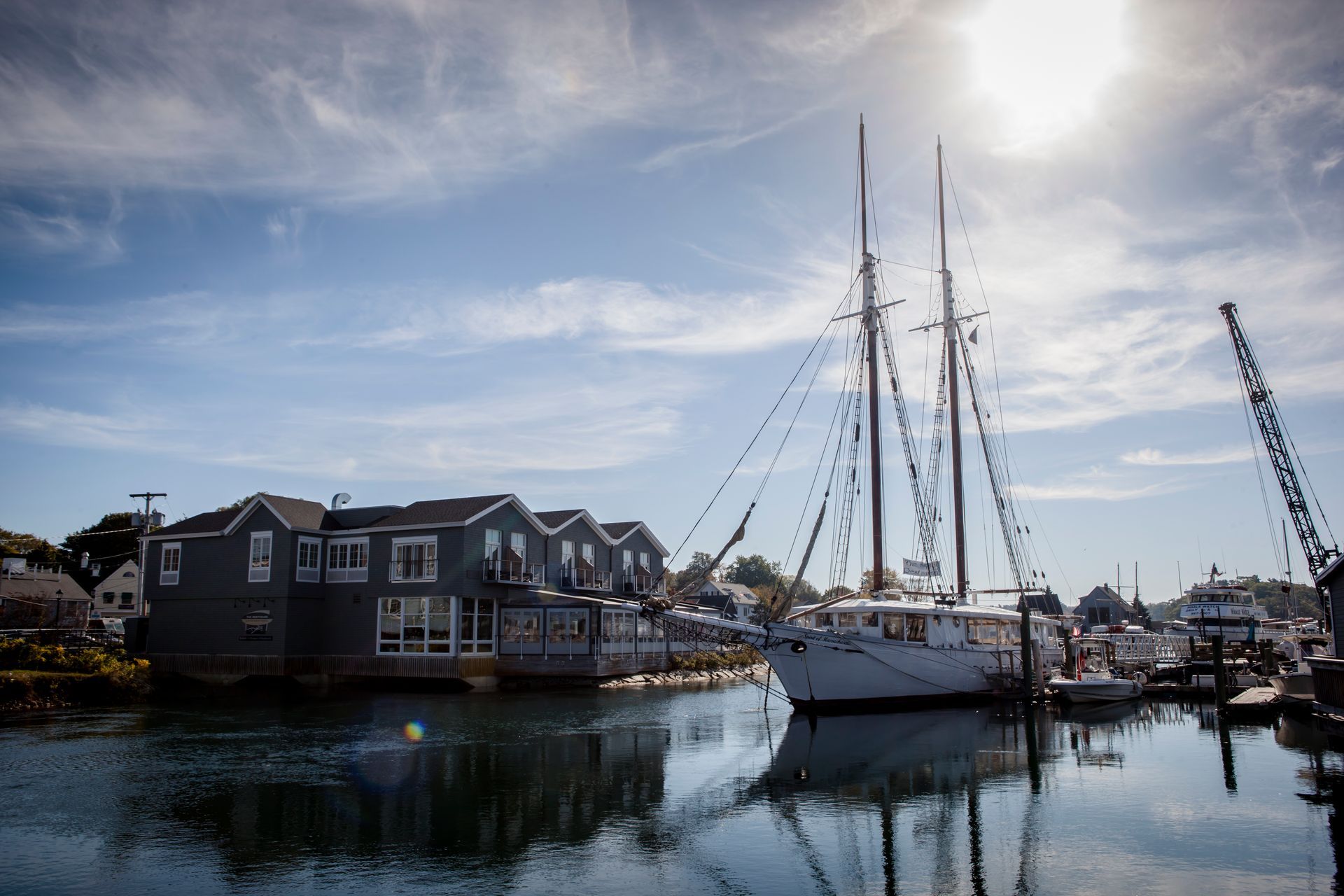 Sailboats docked beside waterfront buildings under a bright sun, reflected in calm harbor water.