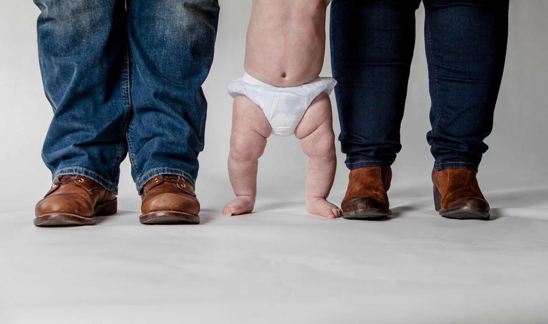 Baby in diaper standing between two adults’ legs on a white floor, wearing blue jeans and brown shoes.