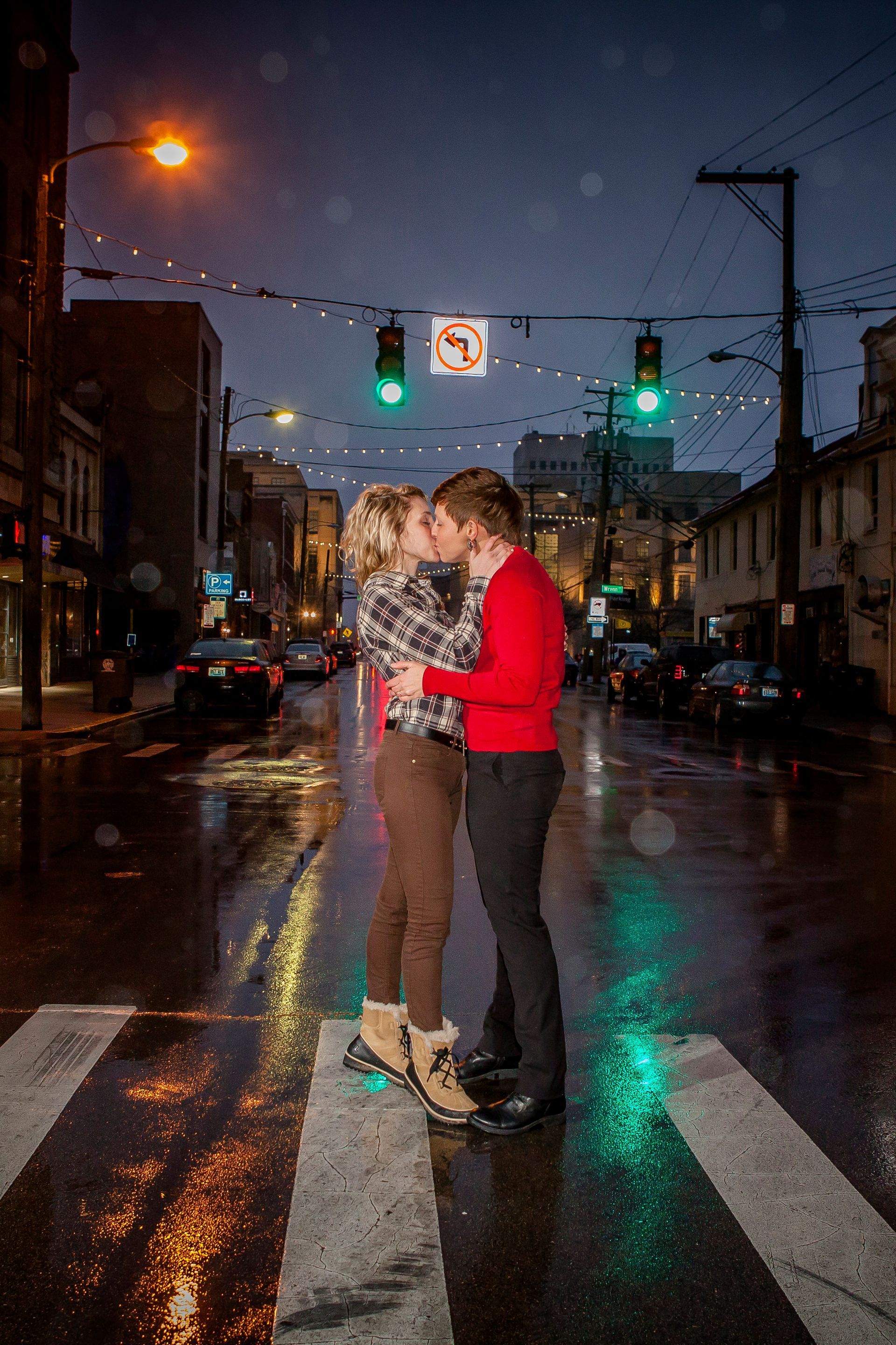 Two people embrace on a wet city street at night under green traffic lights.