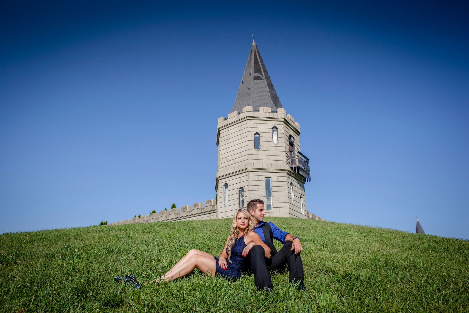 Two people sitting on a grassy hill in front of a stone tower under a clear blue sky