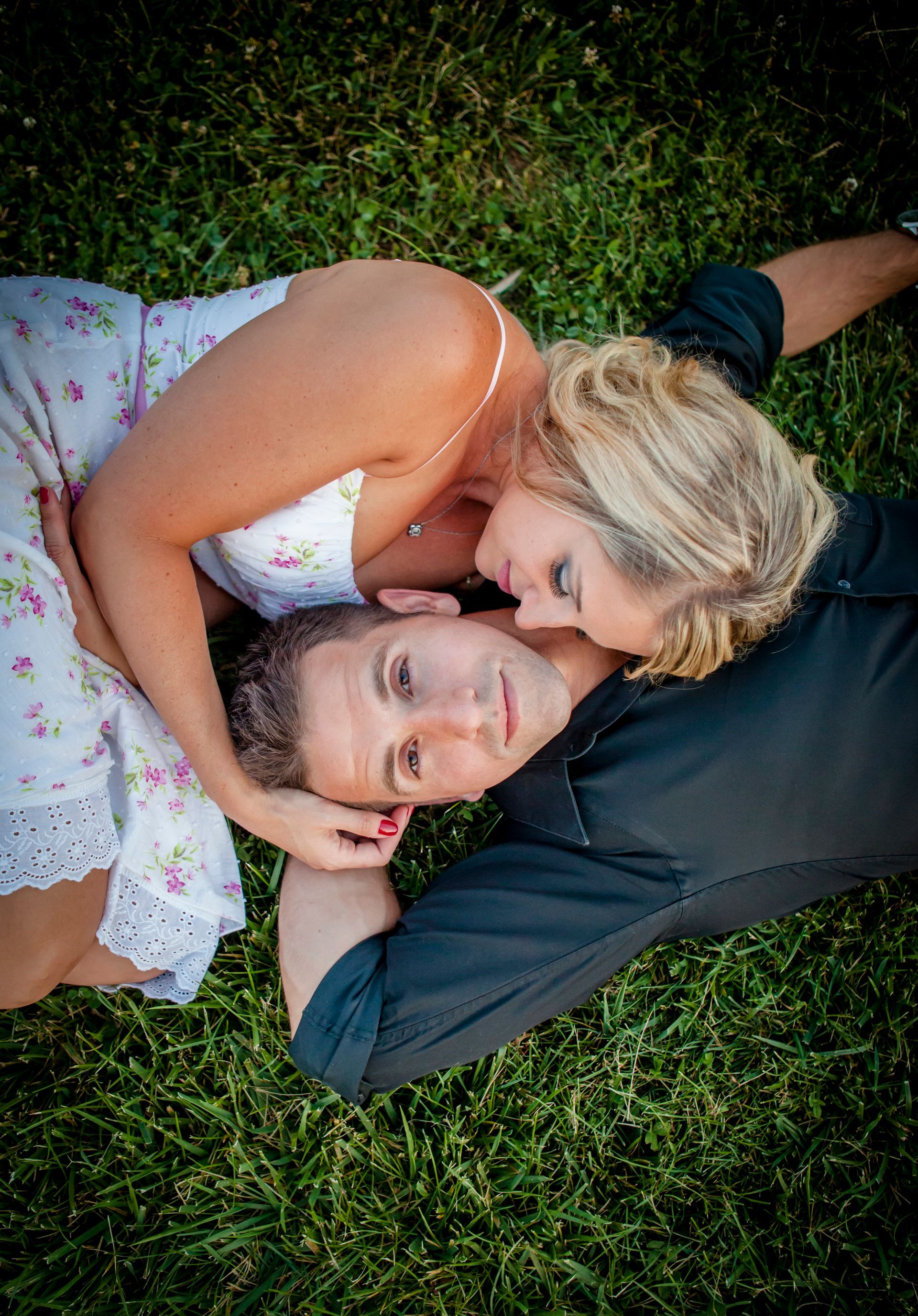 Two people lying on grass, one resting their head on the other's shoulder, both smiling softly.
