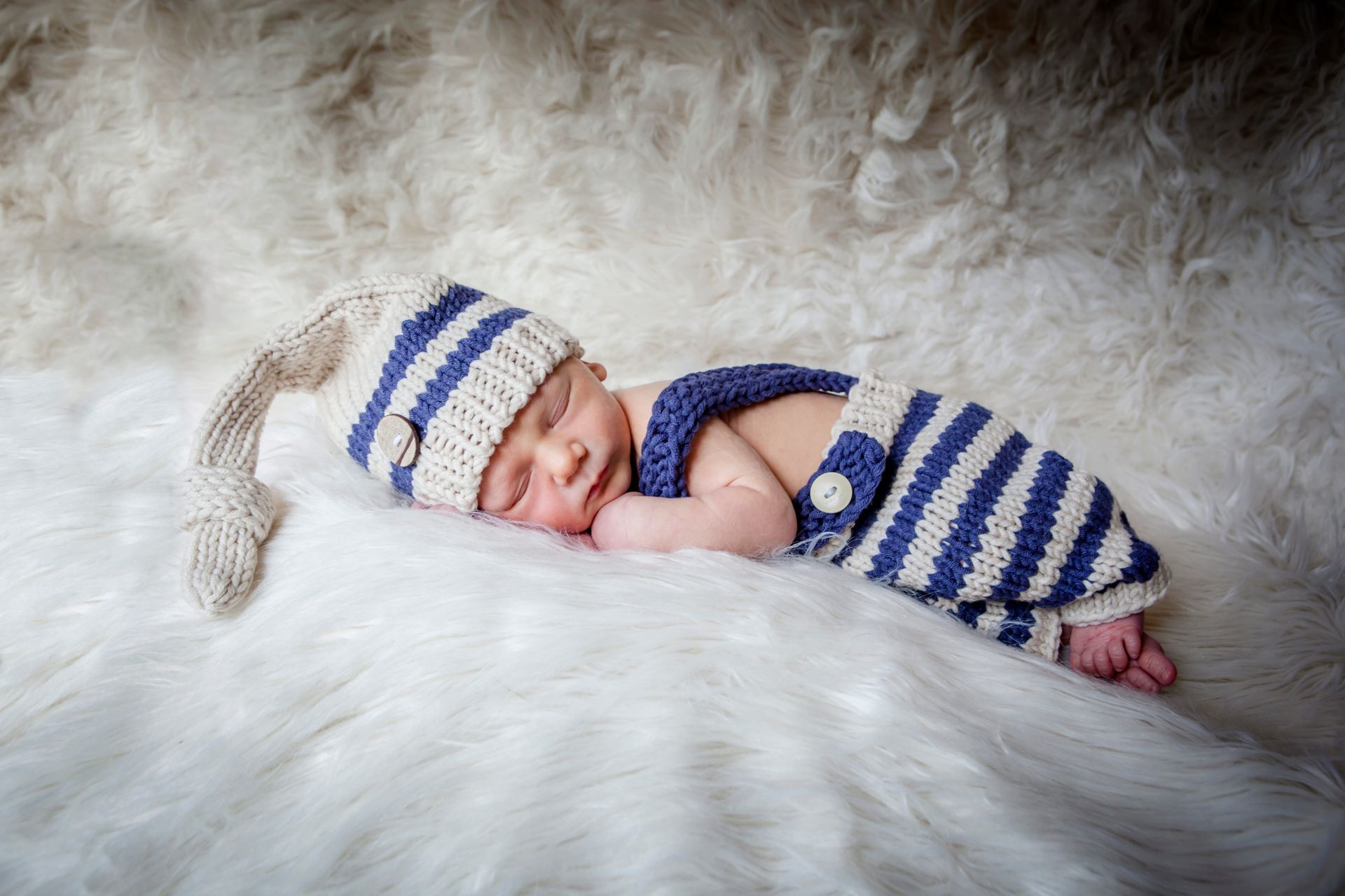 Sleeping newborn in blue-and-white knit outfit and hat on a fluffy white blanket