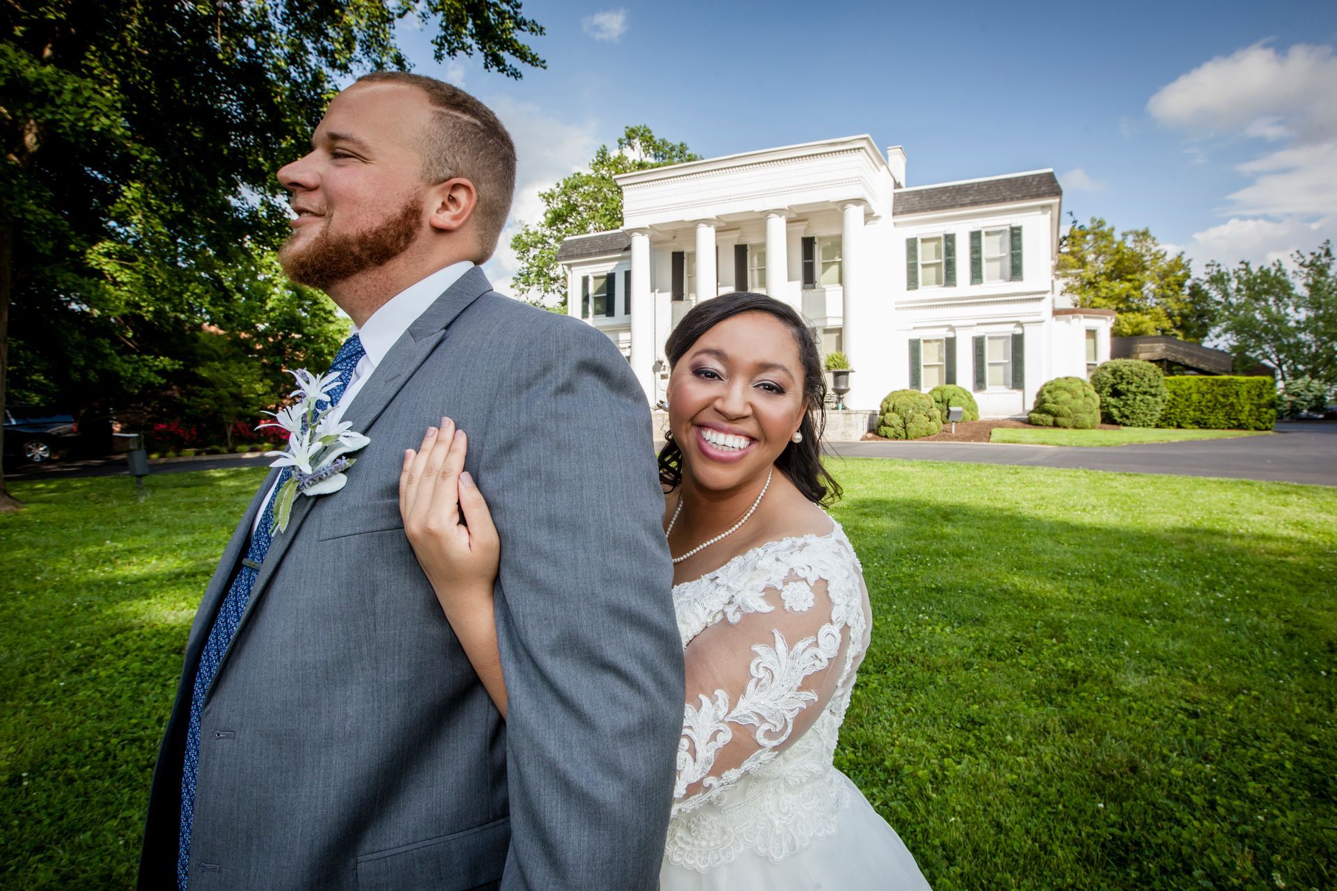 Bride and groom posing on a lawn before a white mansion, smiling back-to-back