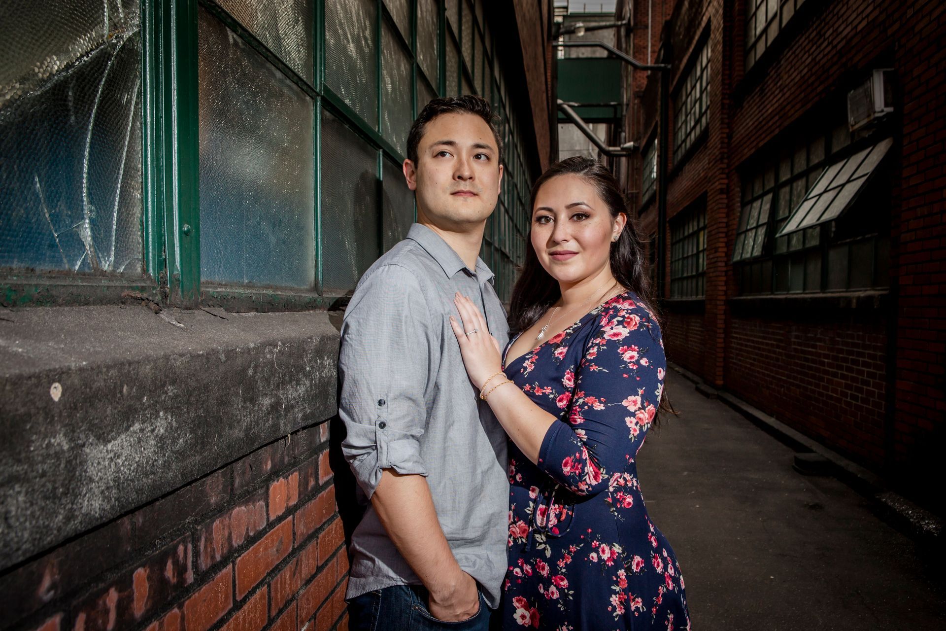 Couple posing in a dim alley, leaning against a brick wall, wearing casual and floral clothing.
