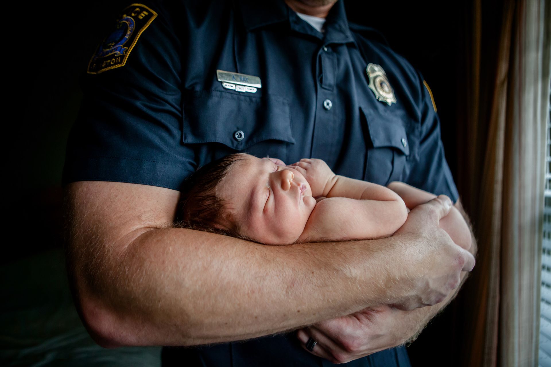 Person in a navy uniform cradles a sleeping newborn wrapped in a blanket near a window
