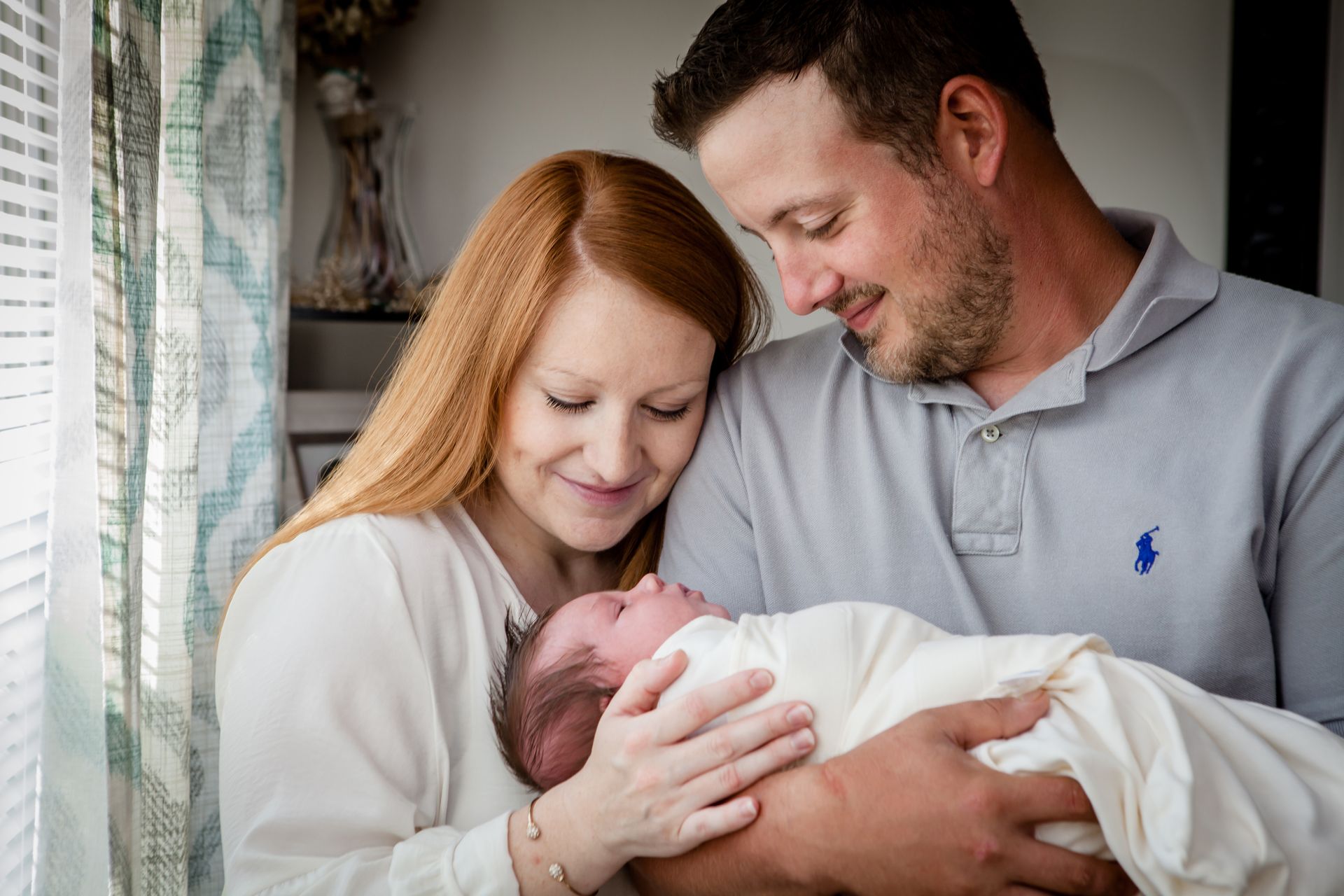 Parents gently holding a newborn by a window, smiling in a soft, cozy indoor setting