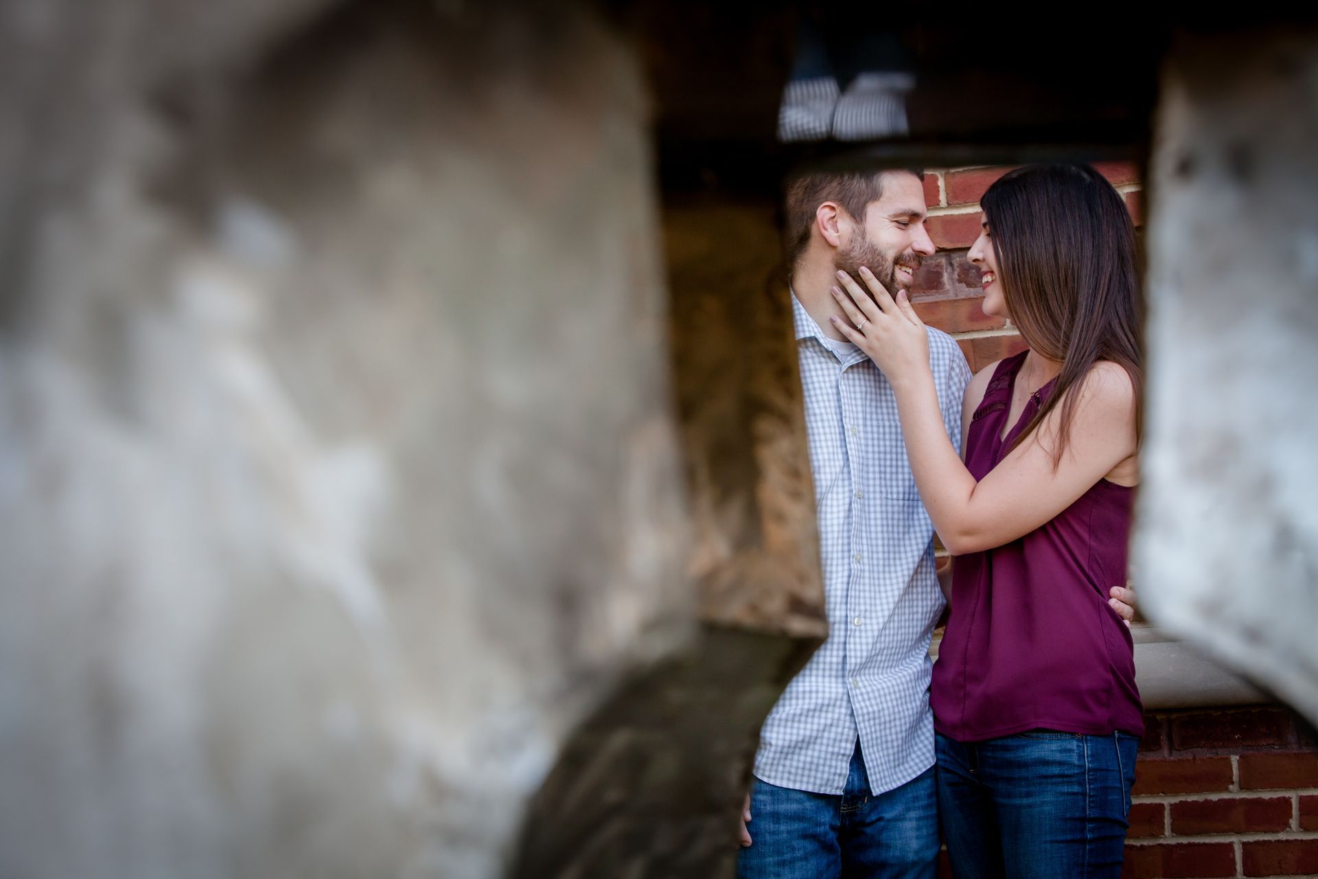 Couple embracing and gazing at each other near a brick wall, framed by a blurred foreground.