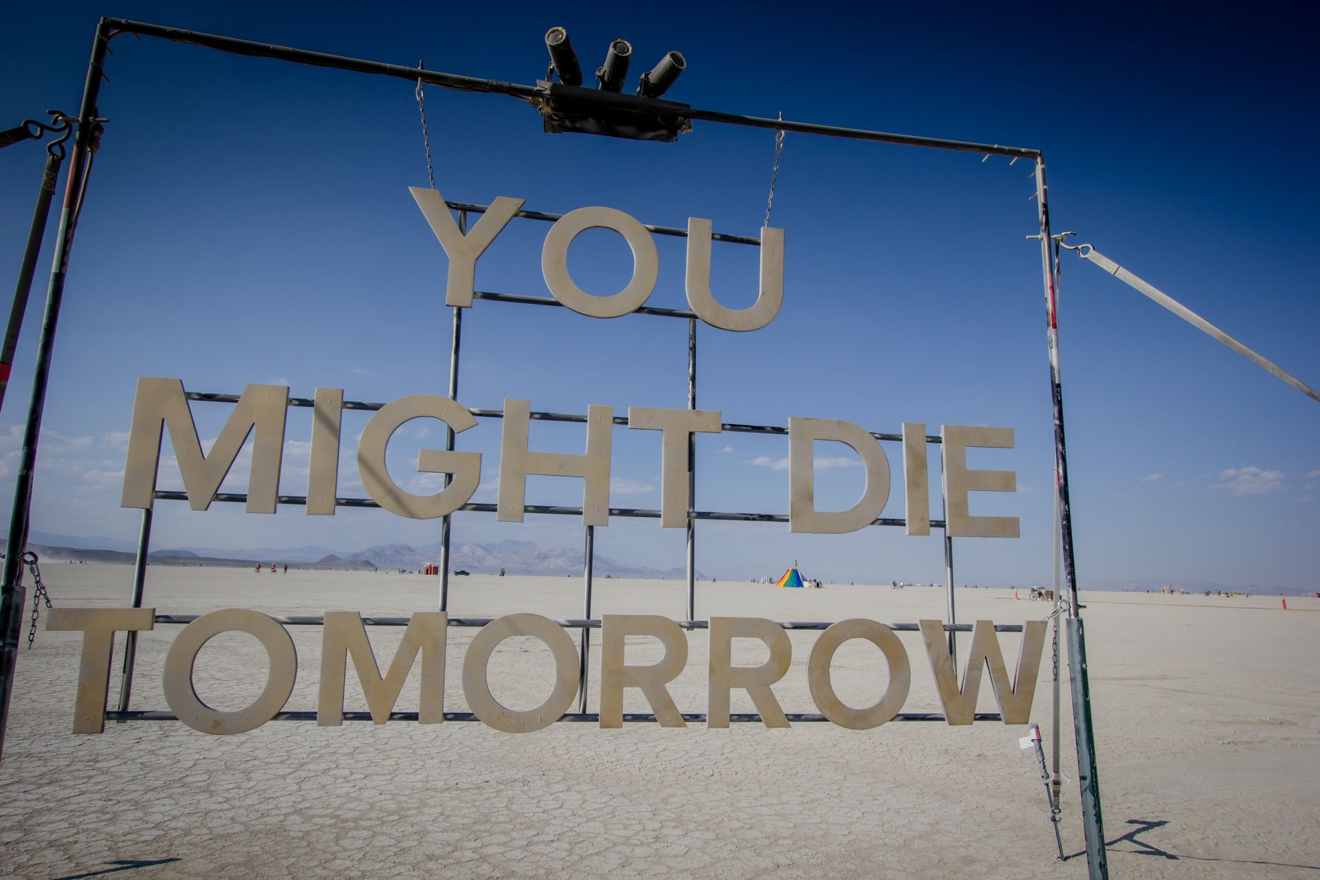 Large sign reading “YOU MIGHT DIE TOMORROW” on a metal frame in a sandy desert under a blue sky