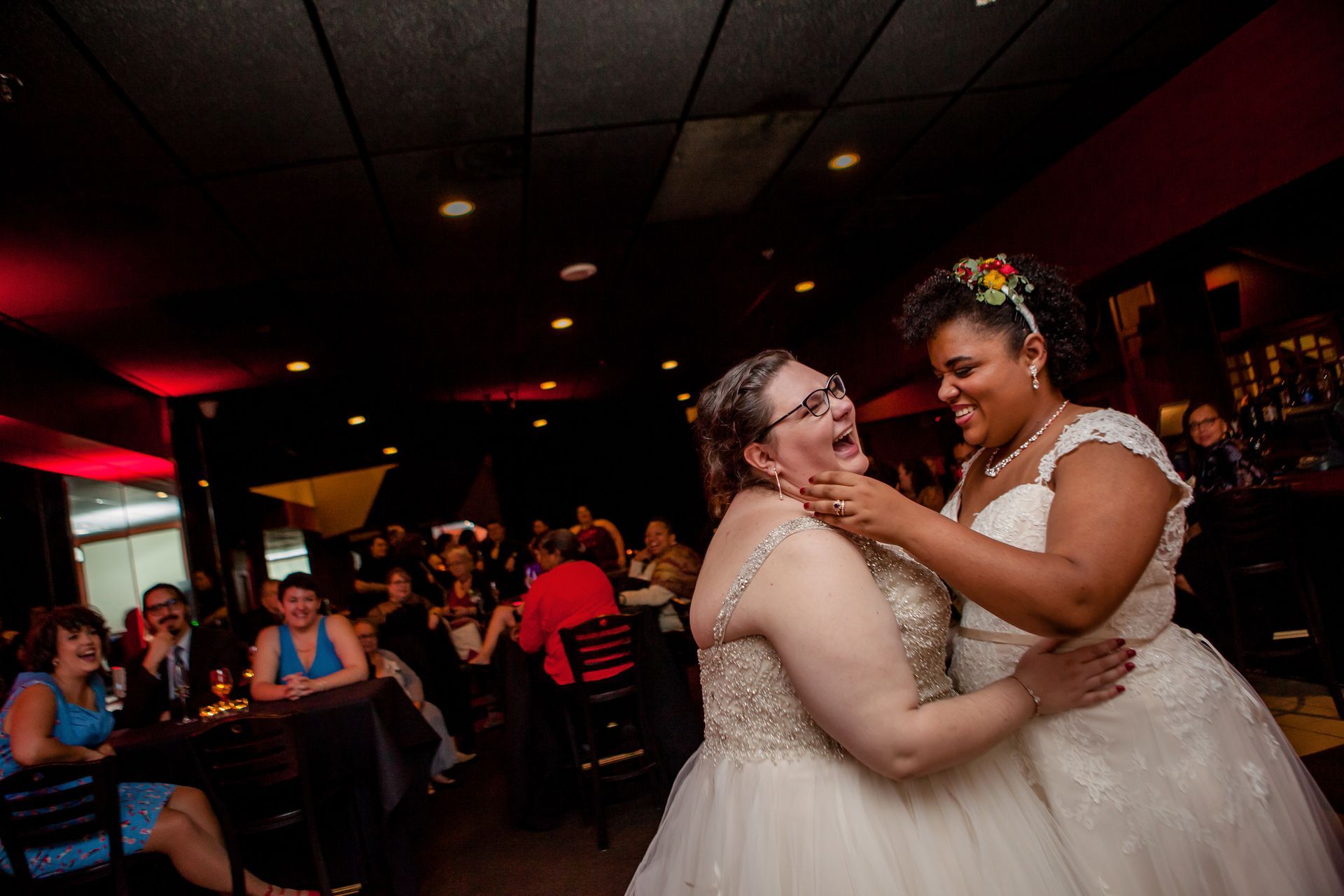 Two brides dancing and smiling at a dimly lit reception, with guests seated in the background.