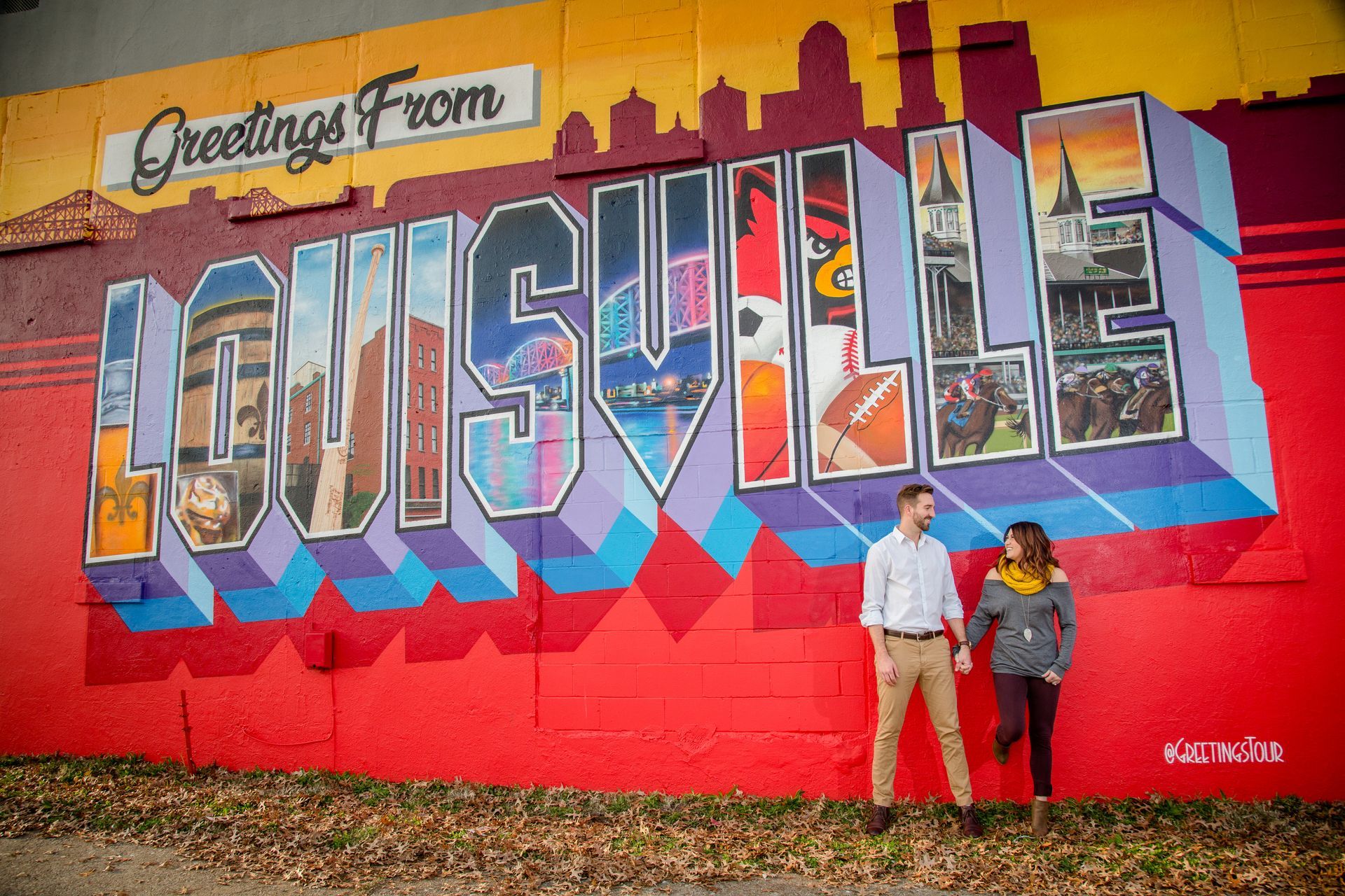 Two people standing before a colorful “Greetings from Louisville” mural.