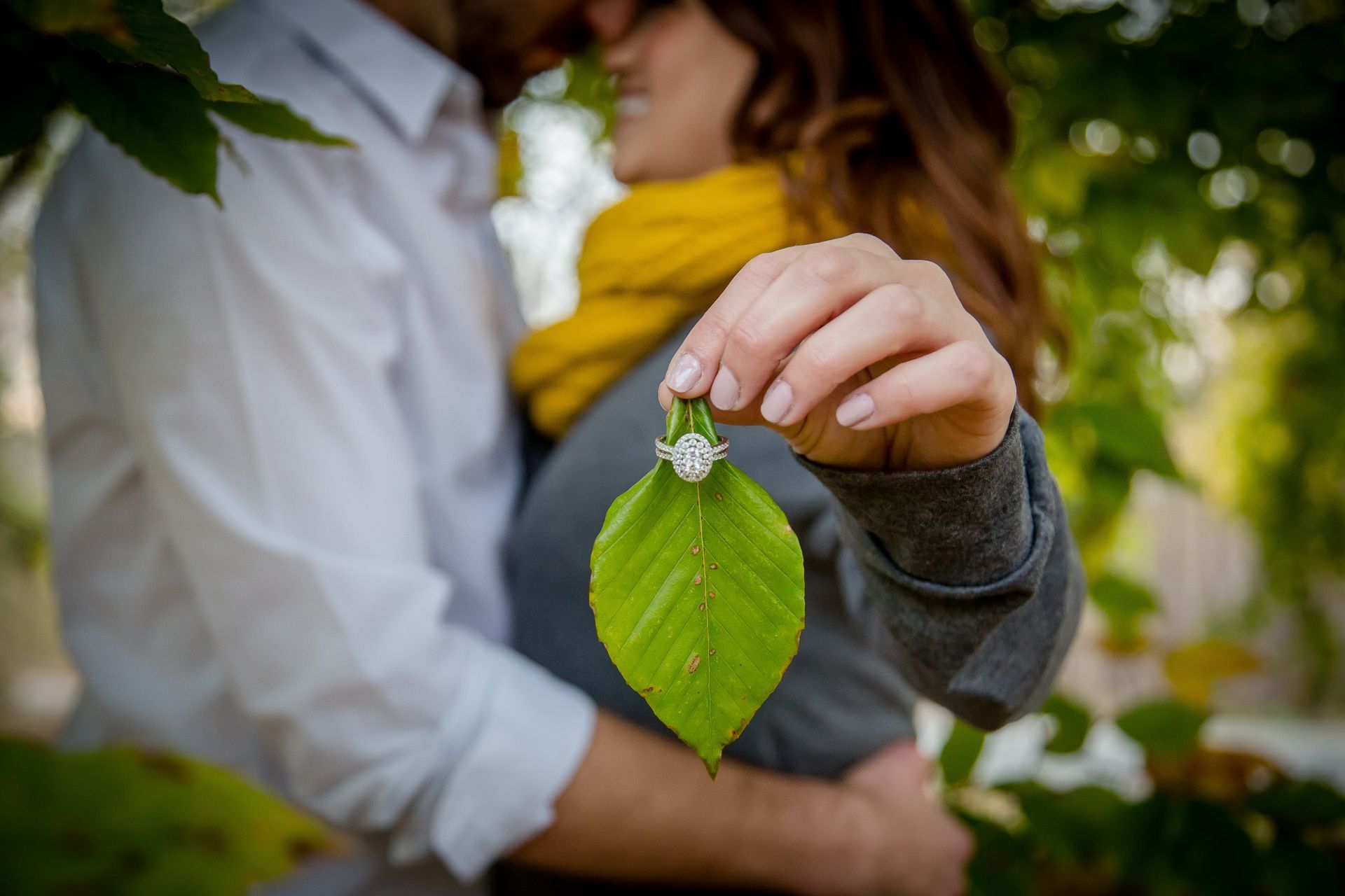 Person holding a green leaf with raindrops in a garden, with another person blurred in the background