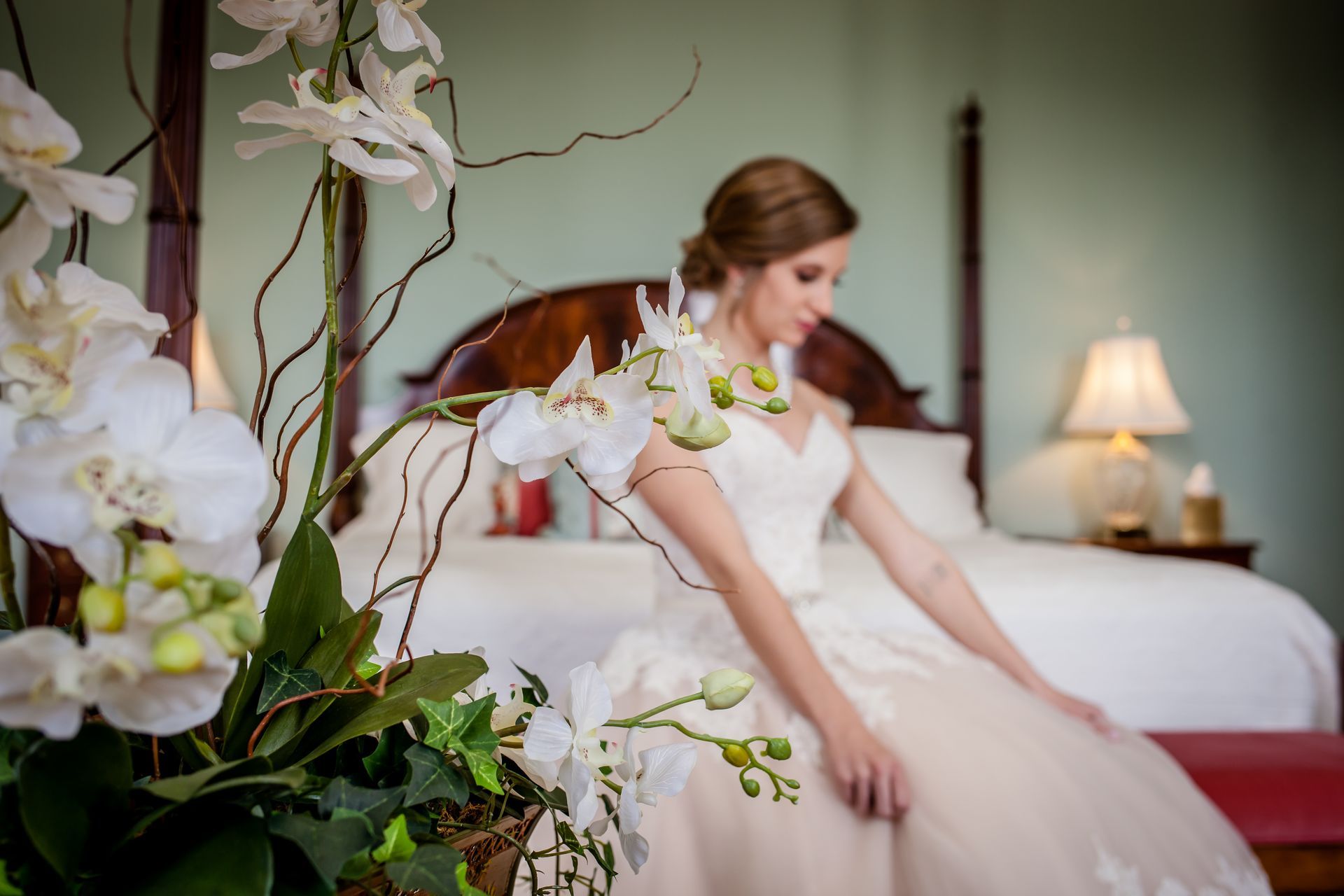 Bride in a white dress sitting on a bed beside white orchids in a softly lit bedroom.