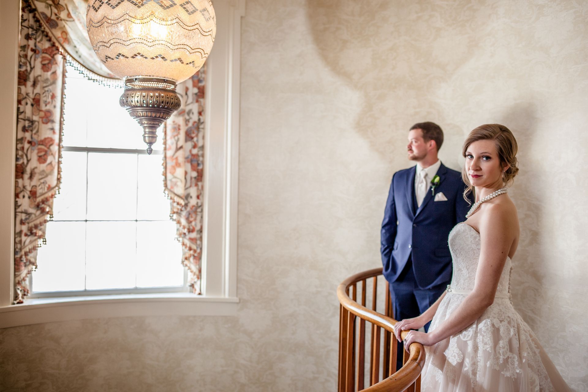 Bride and groom in formal attire by a window with a hanging chandelier in a cream-colored room