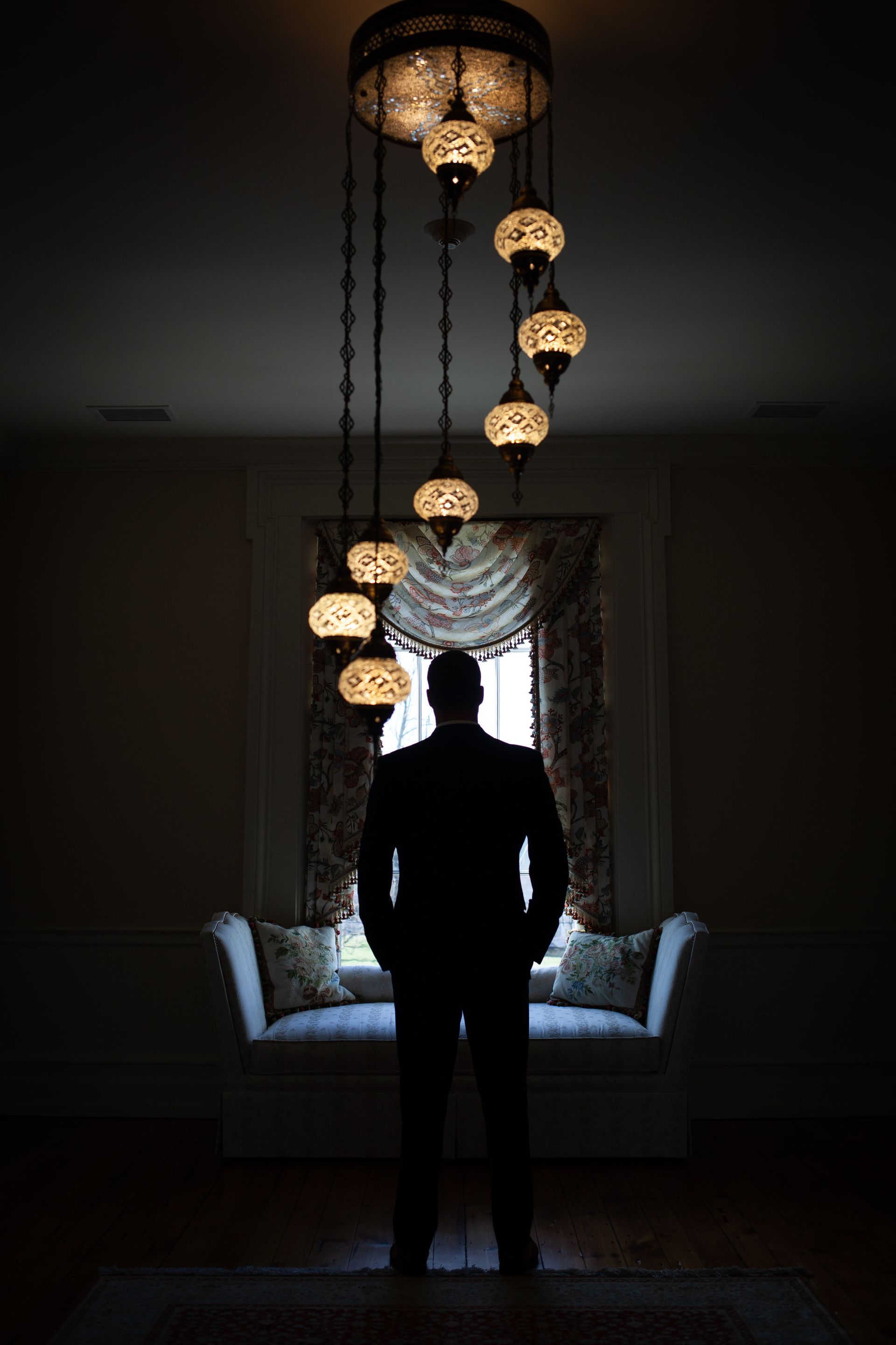 Silhouetted person standing in a dim room beneath hanging pendant lights by a window and sofa