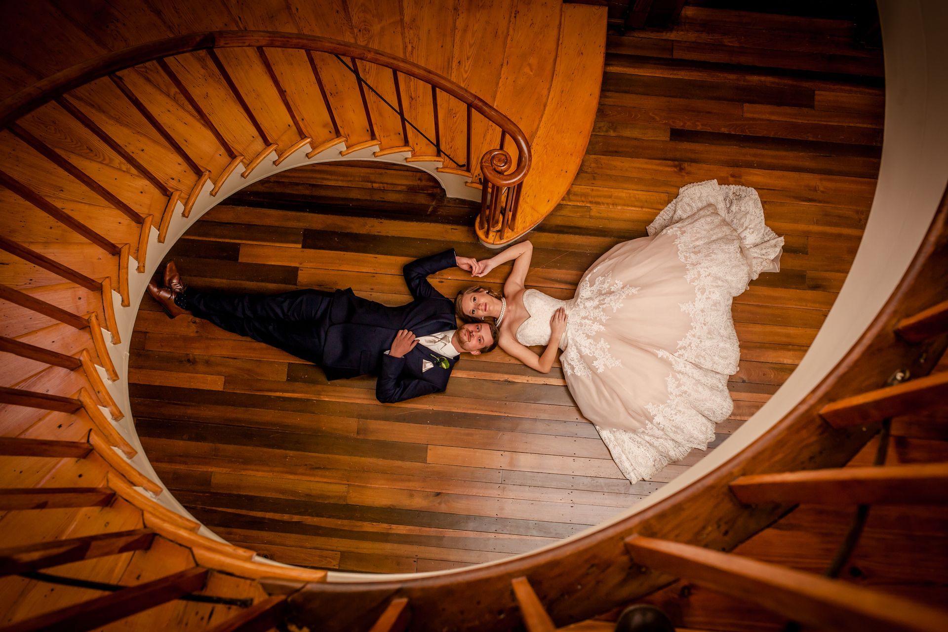 Bride and groom lying on a curved wooden staircase, bride in white gown and groom in black suit