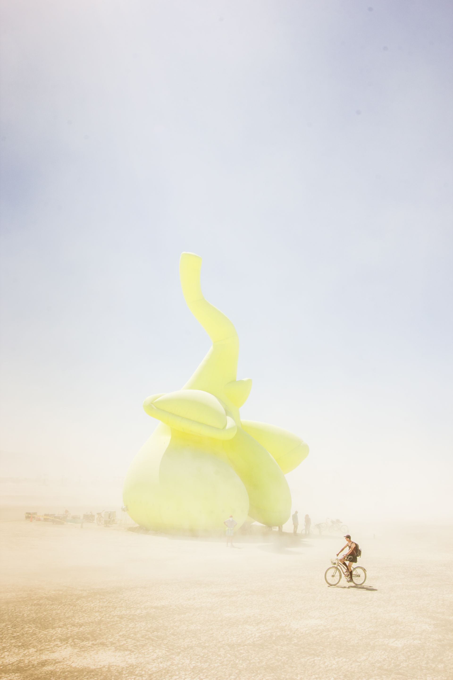 Cyclist riding across a desert toward a giant yellow abstract sculpture under a hazy sky