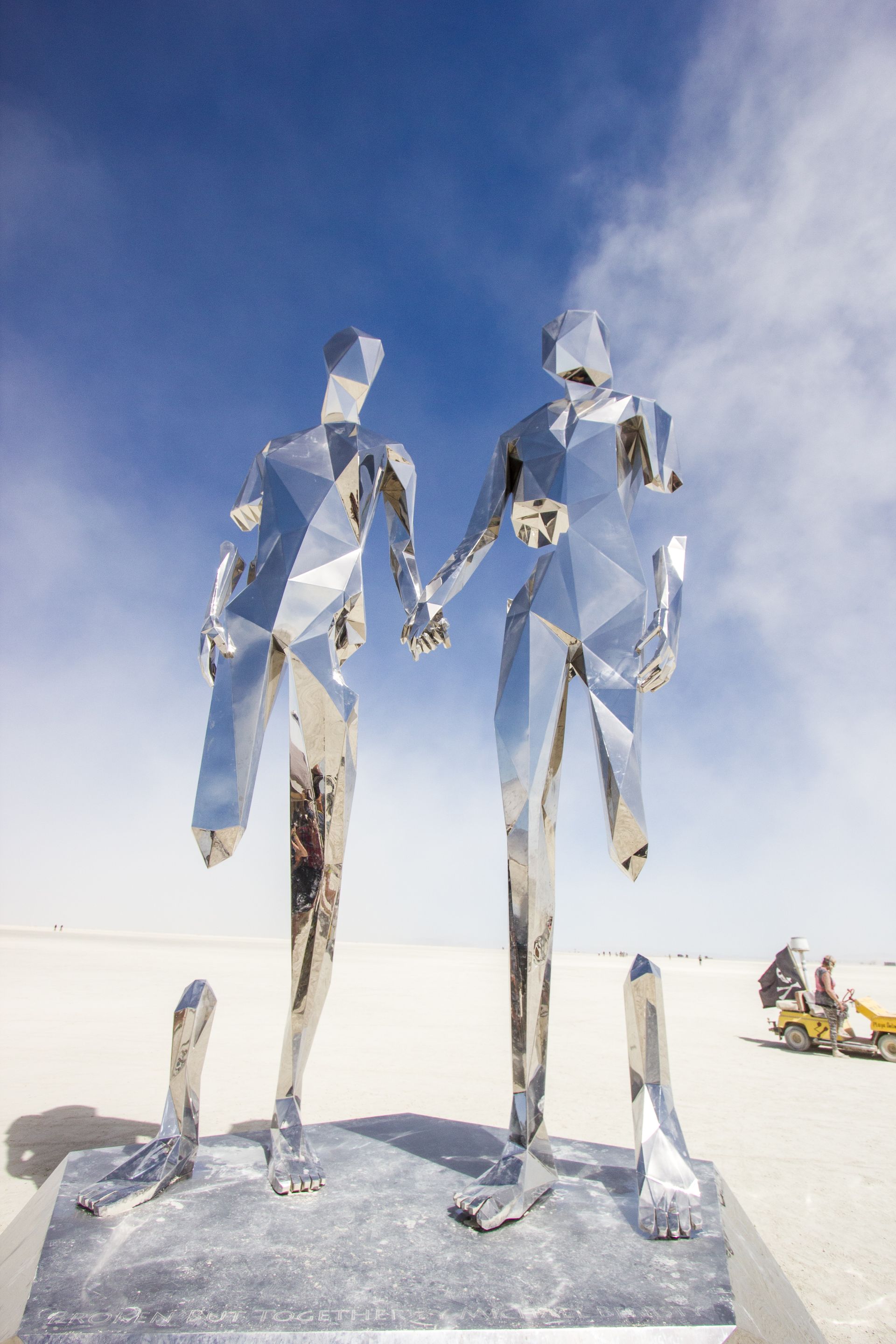 Two mirrored reflective metal sculptures standing on a beach under a blue sky